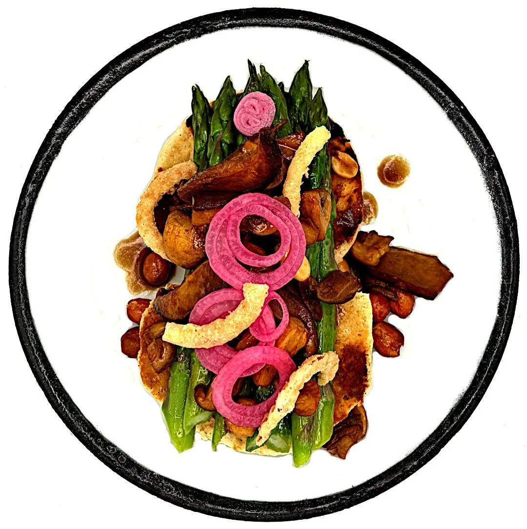 A plated dish with green asparagus spears, pickled red onions, crispy onion strips, roasted mushrooms, and pea hummus on a black-rimmed white plate.