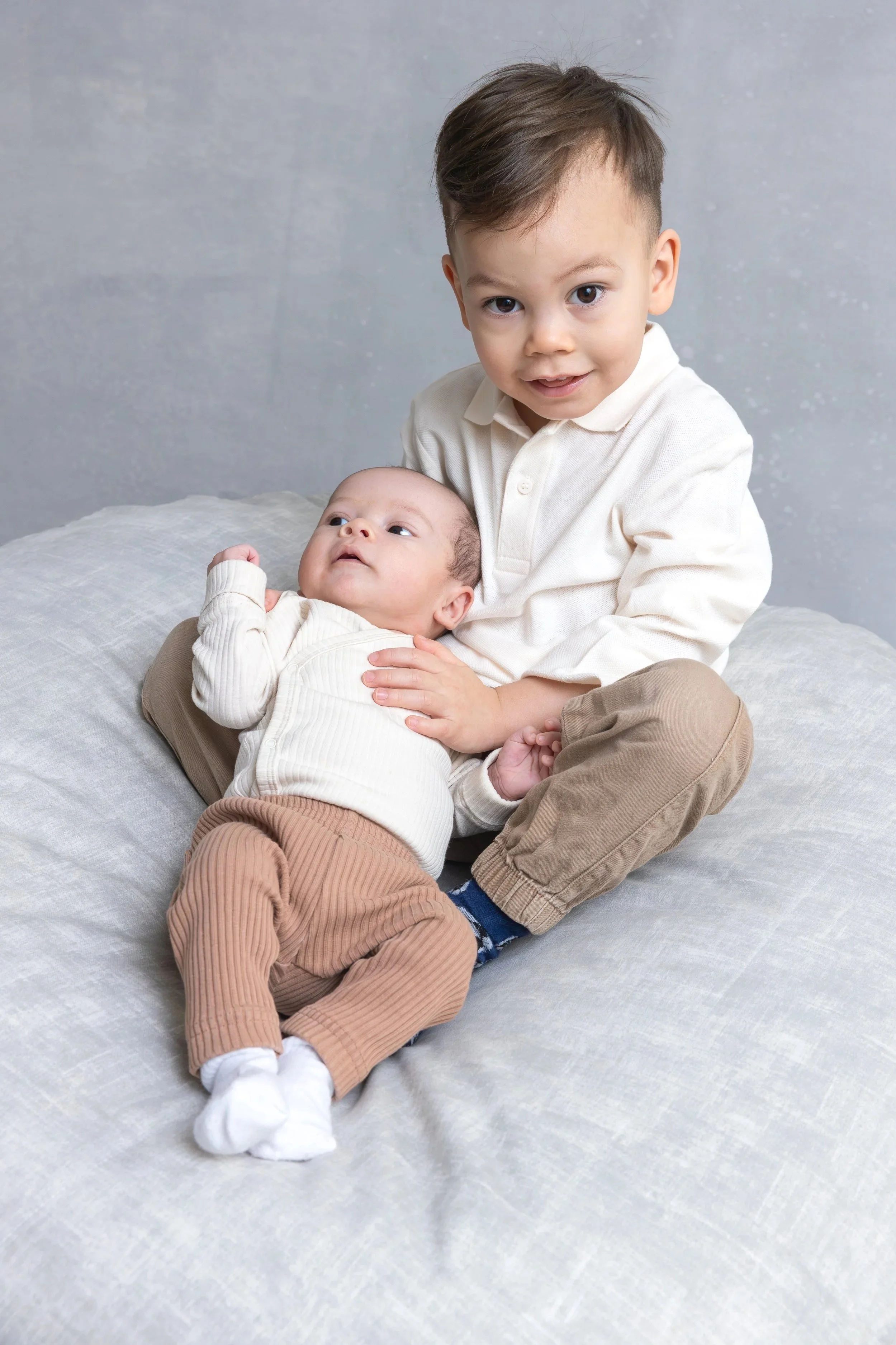 Kuvassa on kaksi lasta, vauva ja toinen pieni lapsi. Vauva makaa isomman lapsen sylissä valokuvastudion säkkituolilla. Lapsilla on vaaleat vaatteet päällään. Isompi lapsi katsoo kameraan ja vauva katsoo muualle.