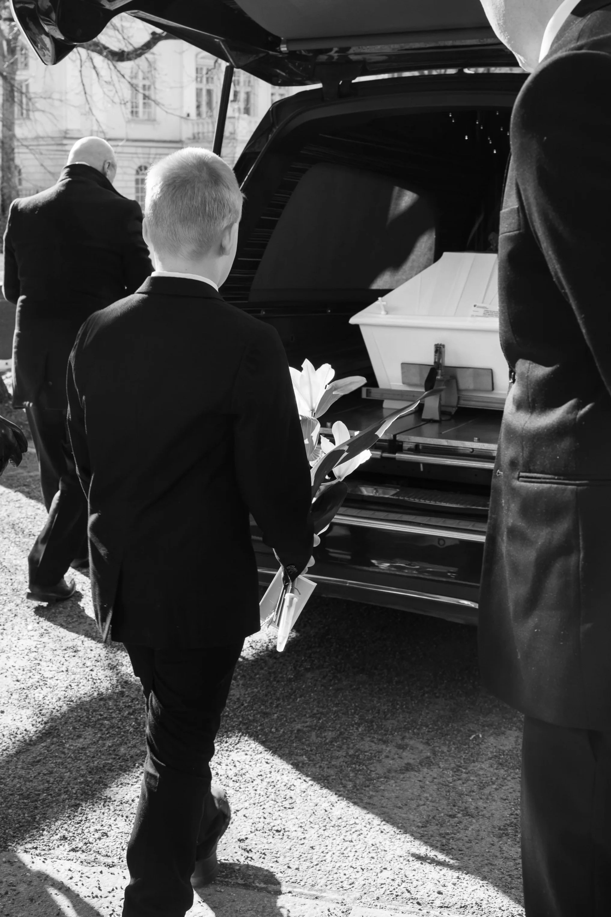 Mustavalkoisessa kuvassa poika on laskemassa kukkaa valkoisen arkun päälle hautajaisissa. Arkku on jo nostettu hautausauton kyytiin. Kuvan etualalla on mies tummassa puvussa. Kuvan on ottanut valokuvaaja Iina Ikävalko.