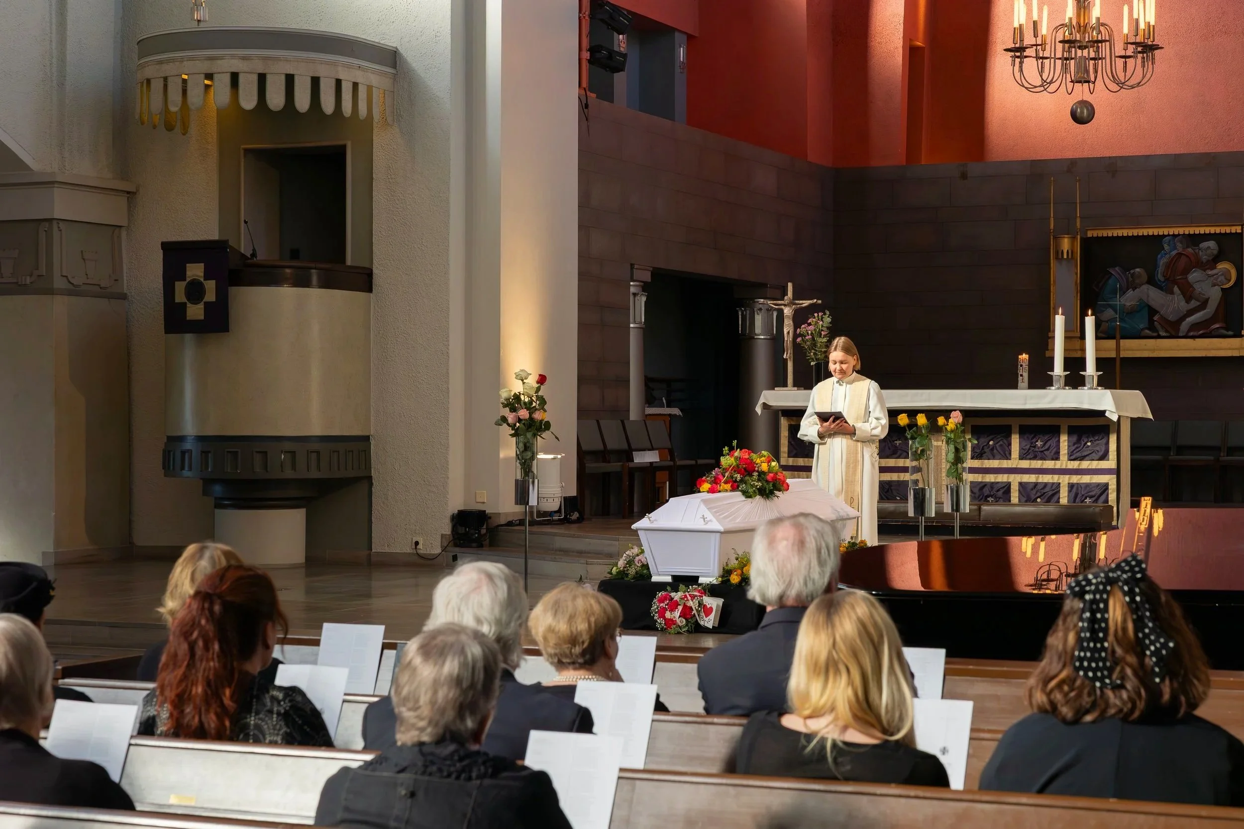 Kuvassa on hautajaiset. Pappi seisoo arkun vierellä pitämässä puhetta. Juhlavieraat istuvat kirkon penkissä Mikael Agricolan kirkossa Helsingissä. Kuvan on ottanut valokuvaaja Iina Ikävalko.