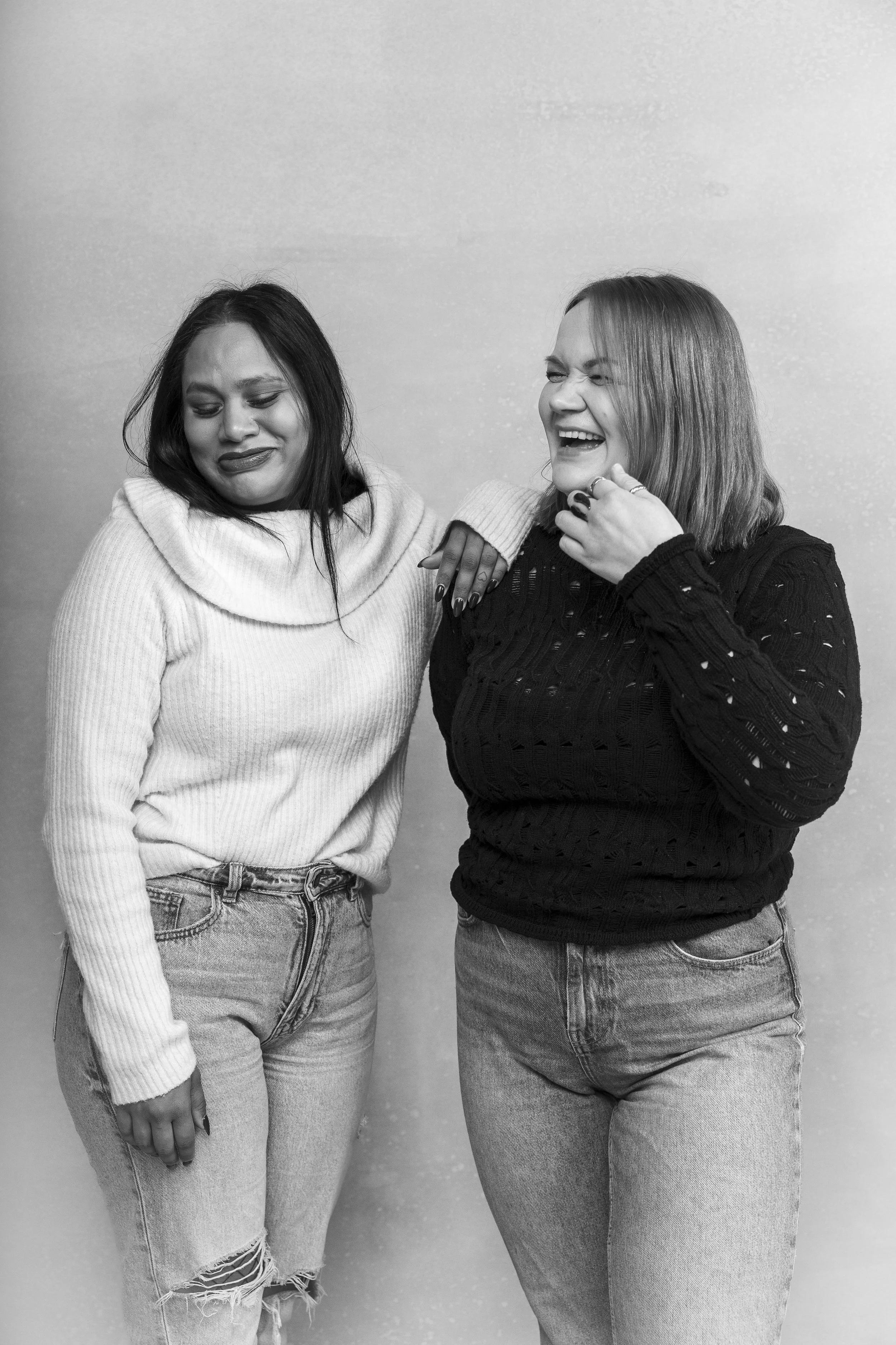 Kuvassa on kaksi naista seisomassa ja nauramassa. He ovat valokuvastudiolla podcastin promokuvauksessa. Valokuva on mustavalkoinen. Naisilla on farkut jalassa ja villapaidat päällään. Valokuvaaja Iina Ikävalko, Helsinki. 