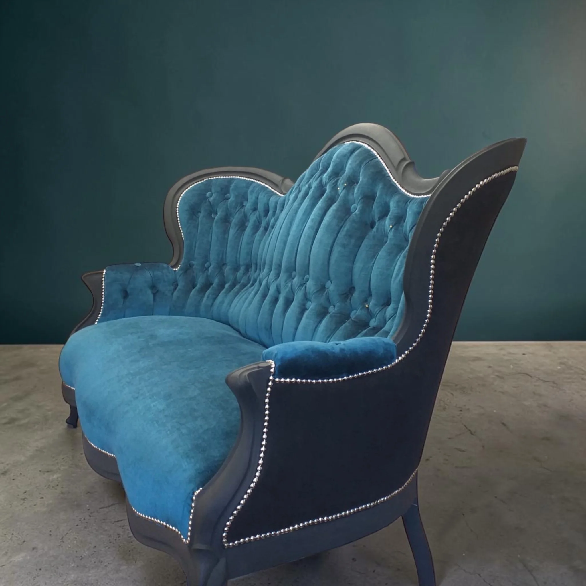 Blue velvet tufted armchair with wooden frame and nailhead trim, placed on a concrete floor against a teal wall.