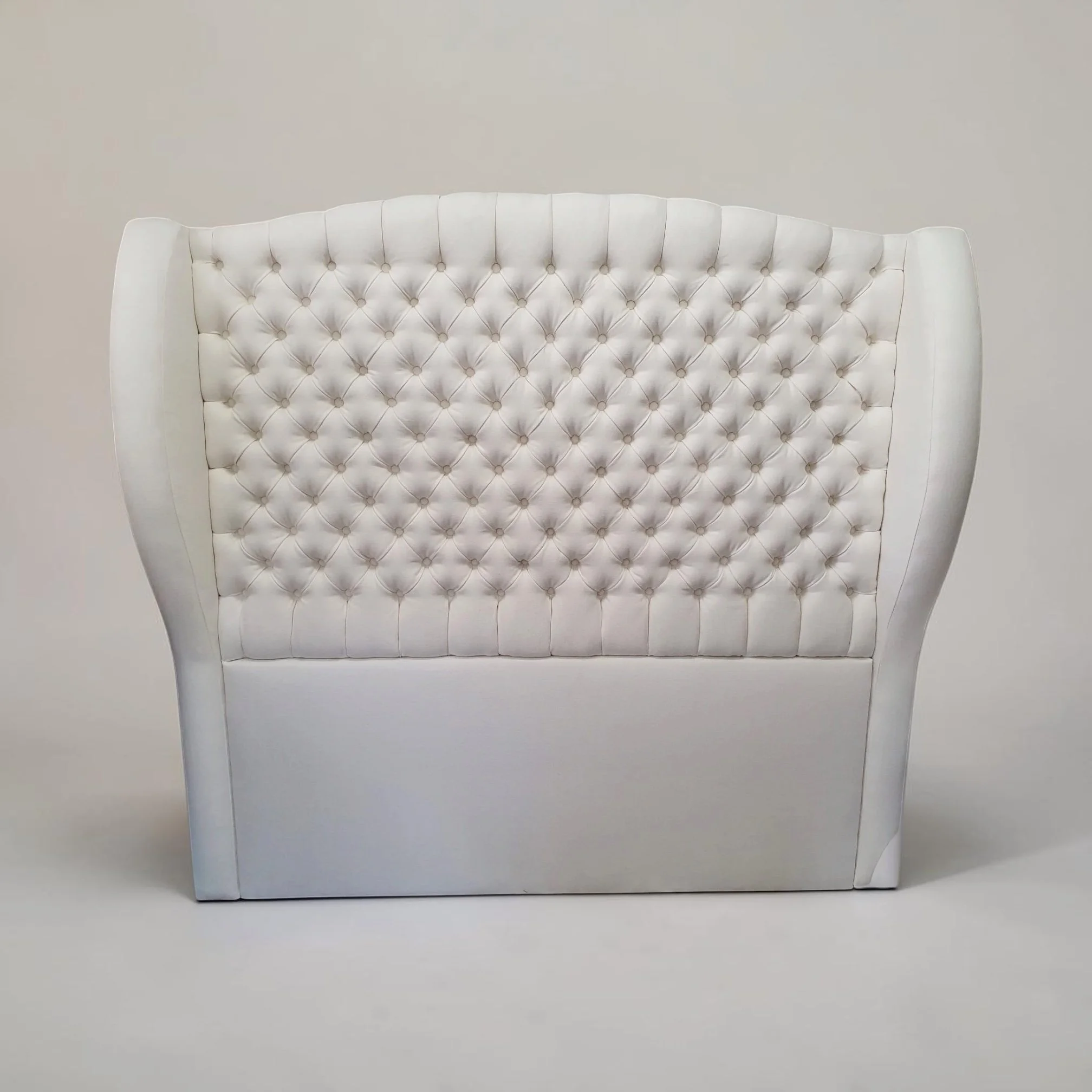 A white, tufted headboard with a curved top edge and button detailing, set against a plain light wall.