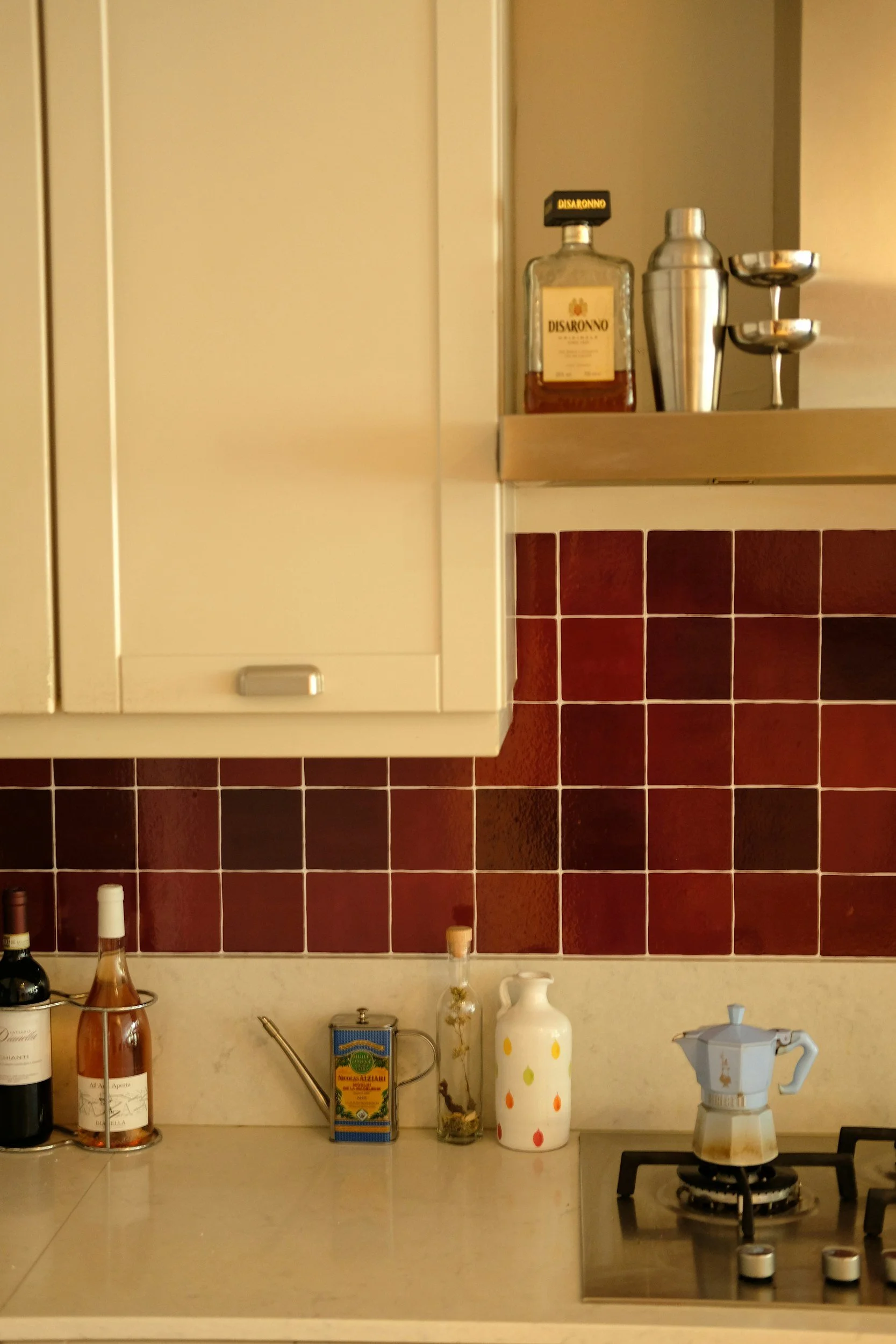 Kitchen countertop with various bottles, containers, and appliances, including a stove with a moka pot, against a background of red tiles and a beige wall with a cabinet.