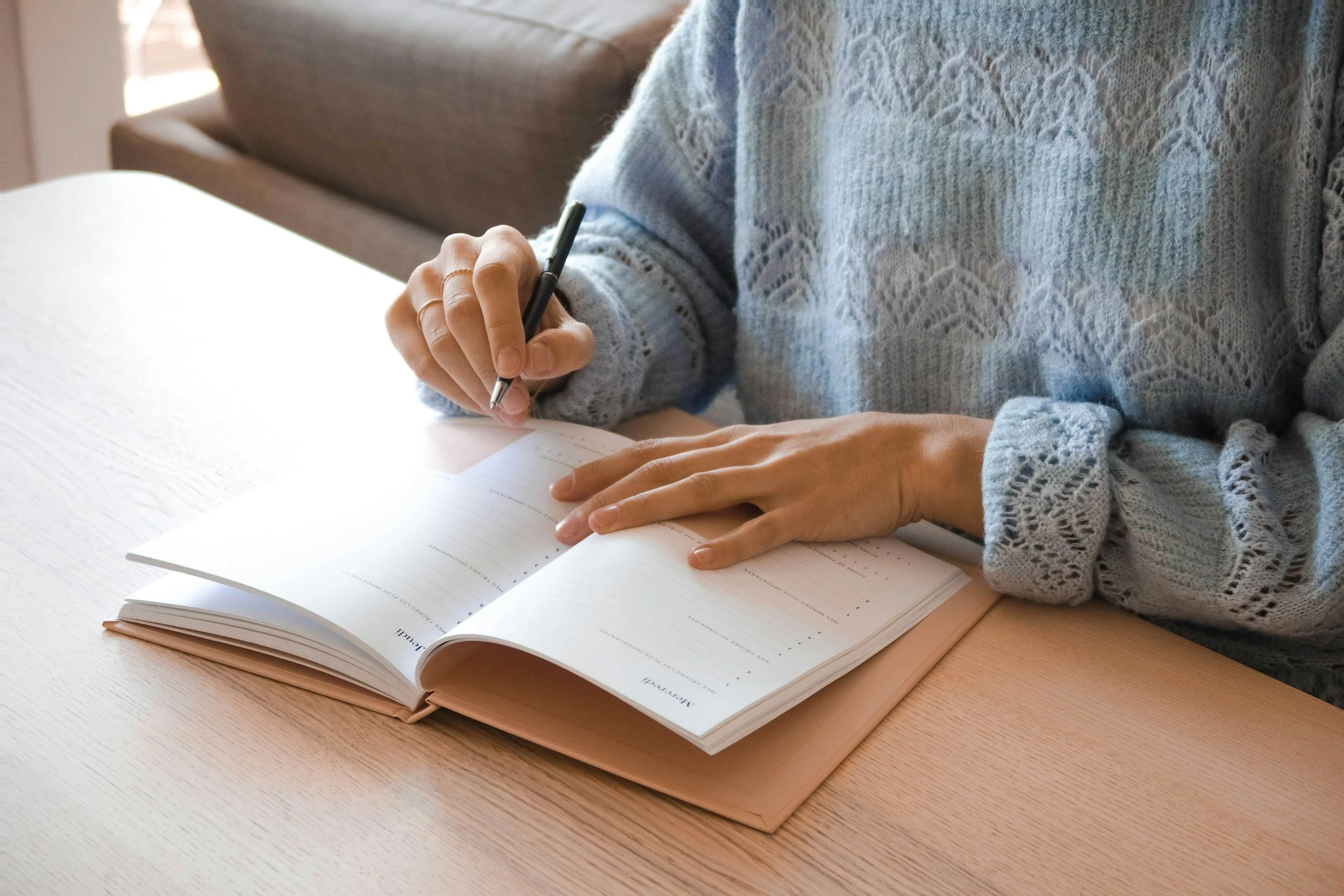 A person wearing a light blue knitted sweater is sitting at a wooden table, writing in a planner or notebook with a black pen. The planner is open, and the person is focused on their writing.