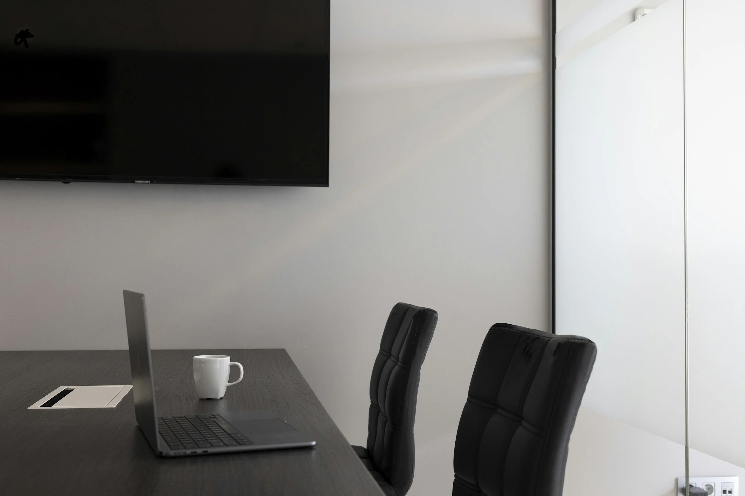 A modern conference room with black chairs around a dark wood table. A laptop and a white coffee mug are on the table, and a large flat-screen TV is mounted on the wall. Sunlight streams through the window, creating bright reflections.