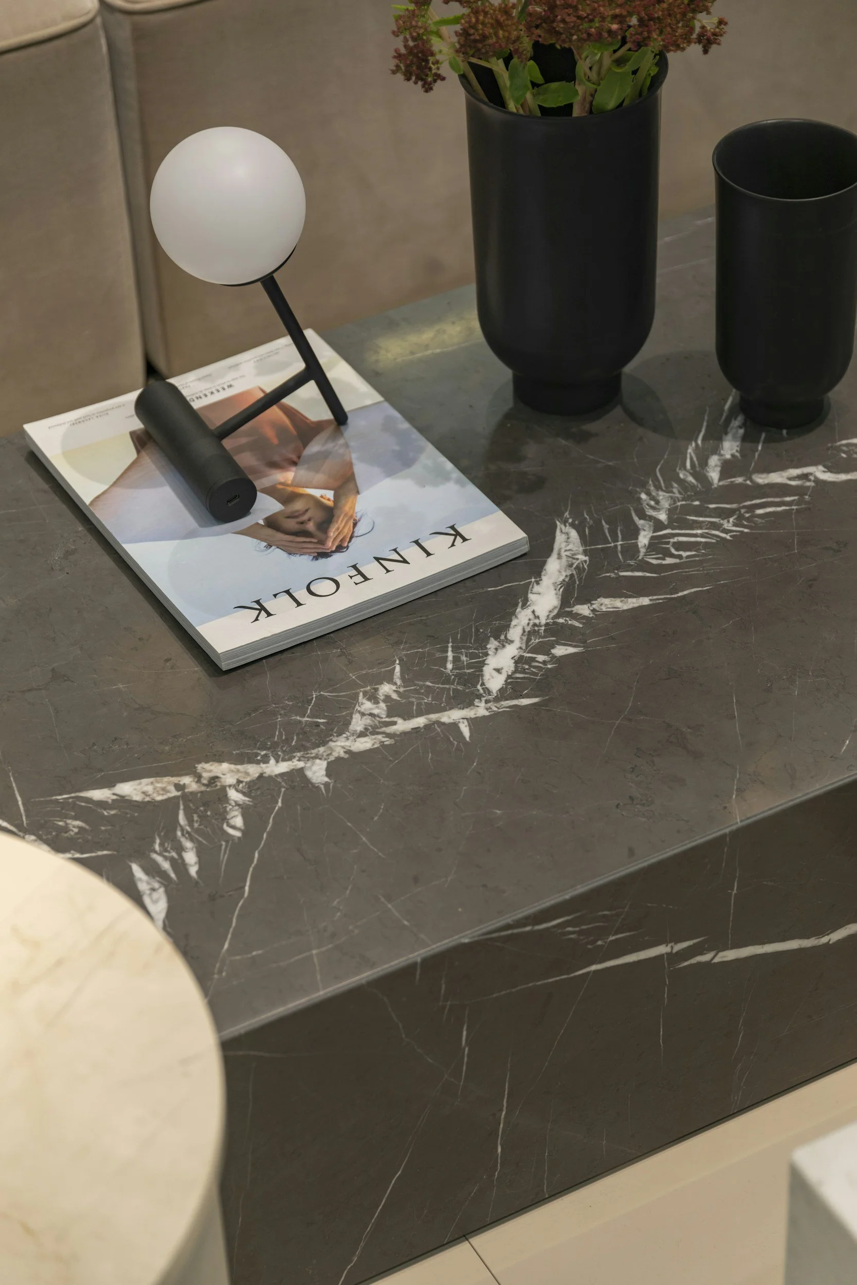 A dark marble table with white veining, holding a magazine, a modern table lamp, and two black vases with a green plant. The background includes a beige couch.