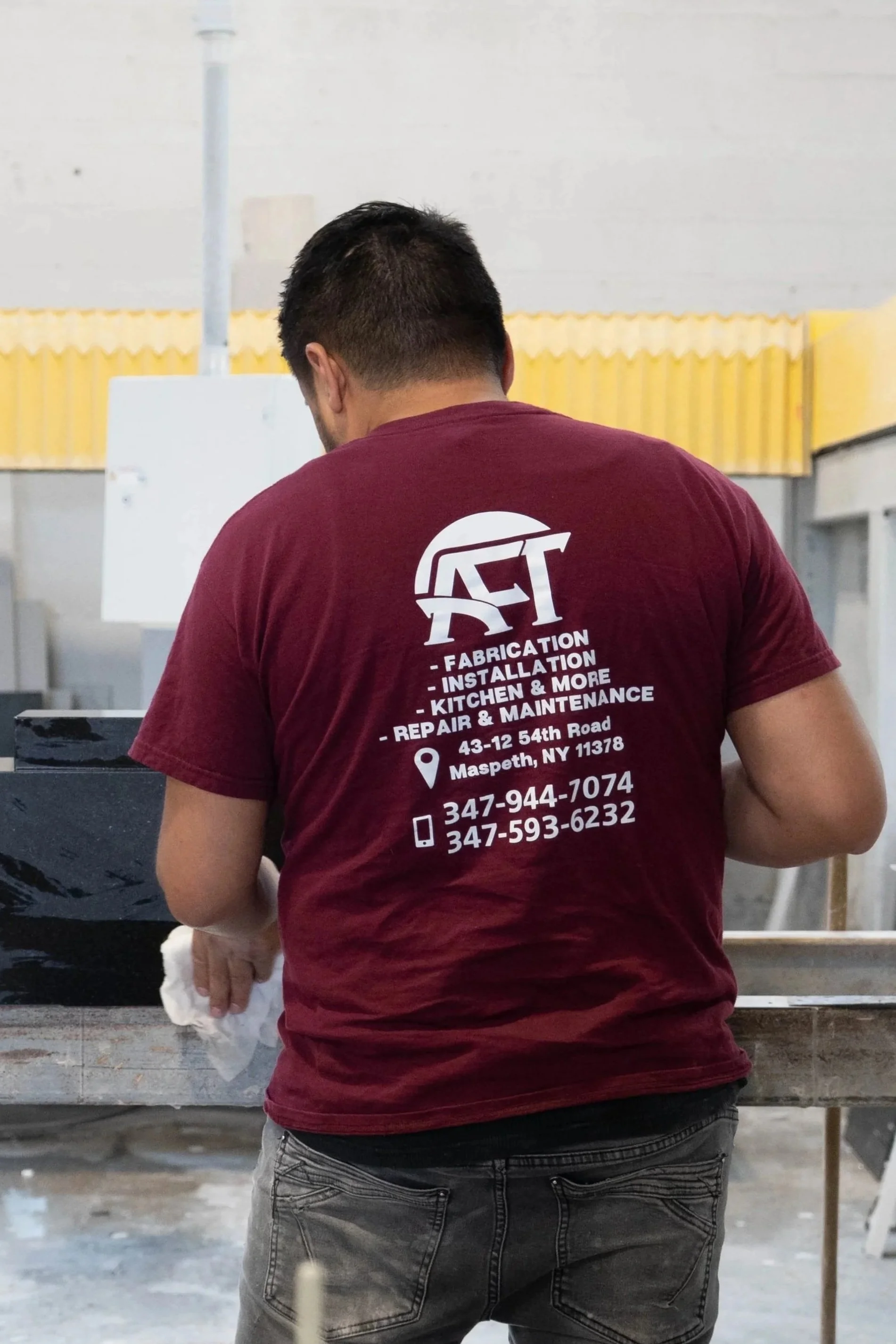Man wearing a maroon T-shirt with a company logo and contact information, working at a construction site or workshop.