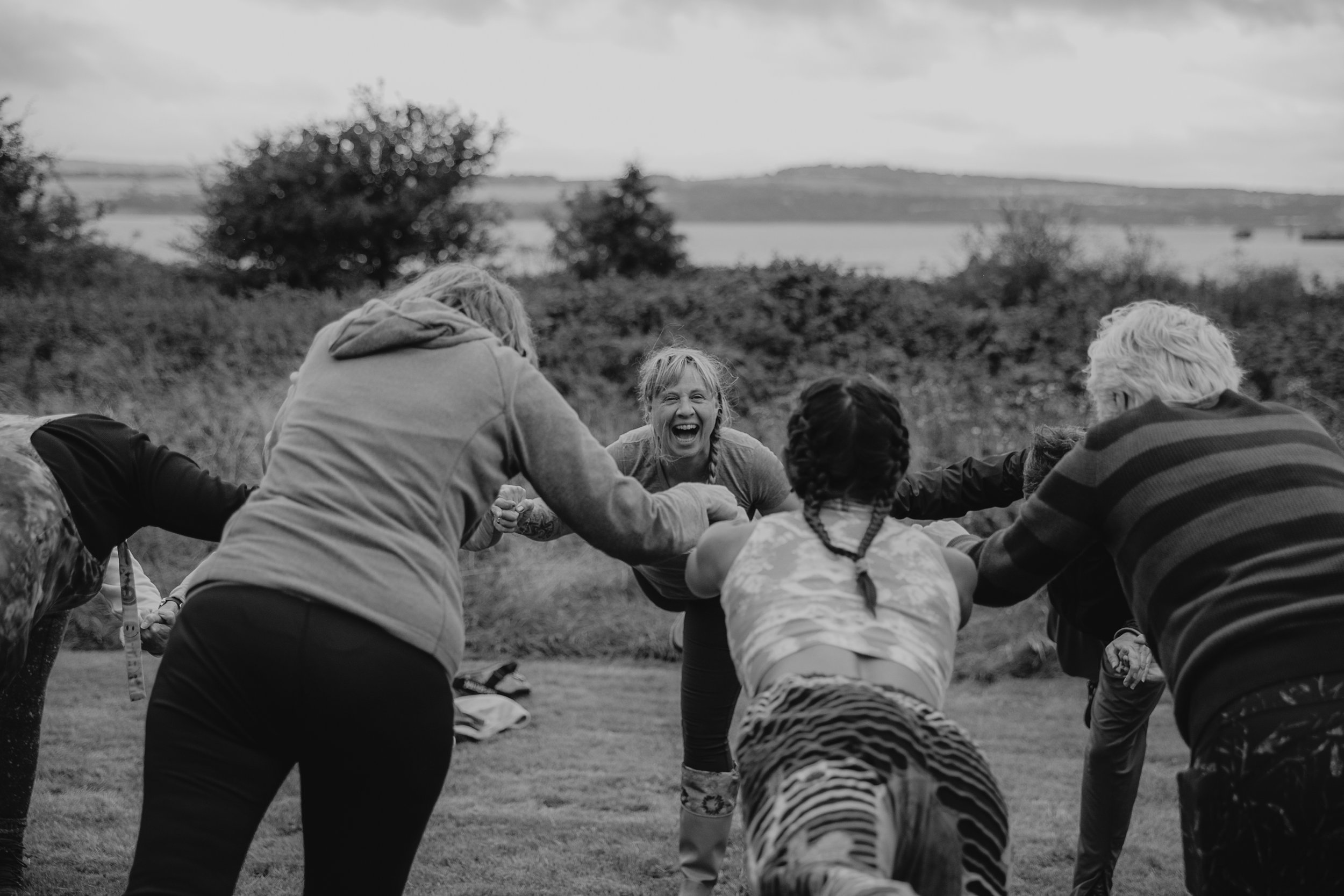Black-and-white photograph of Alice laughing with a group of people practising yoga outdoors, capturing a joyful moment of movement and connection.