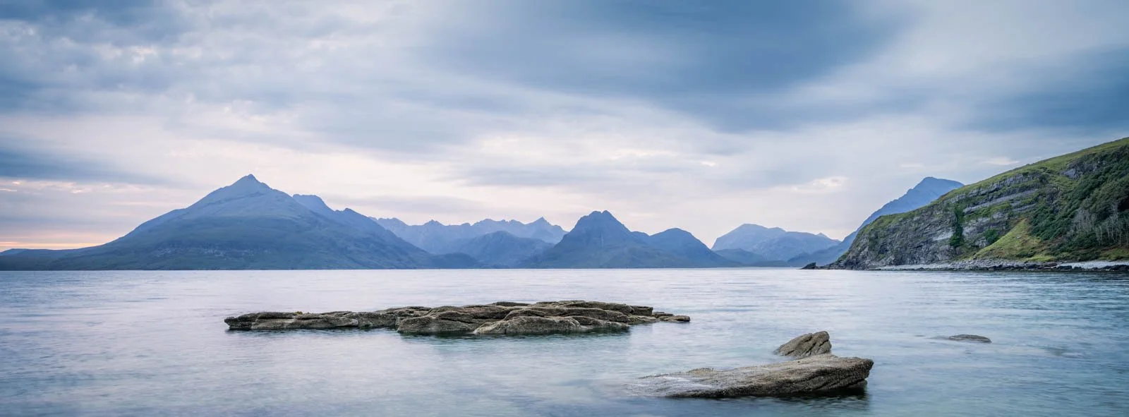 A tranquil lake surrounded by mountains under a cloudy sky, with rocks in the water in the foreground.