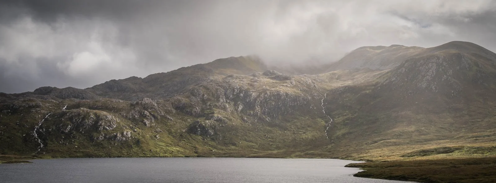 A mountain landscape with a lake in the foreground, steep hills with rocky terrain and small waterfalls, cloudy sky.