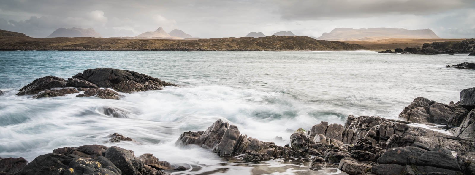 Waves crashing against dark rocks along a coast, with distant mountains under a cloudy sky in the background.