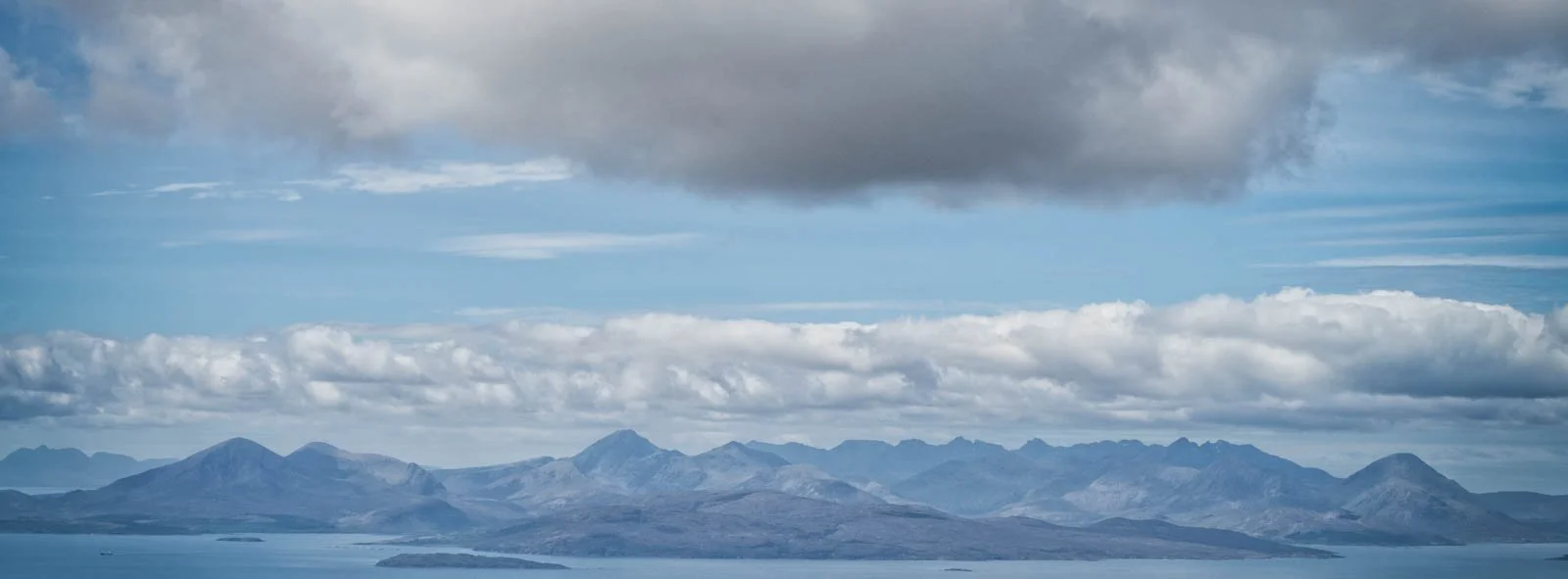 A landscape view of a mountain range with a body of water in the foreground, under a partly cloudy sky.