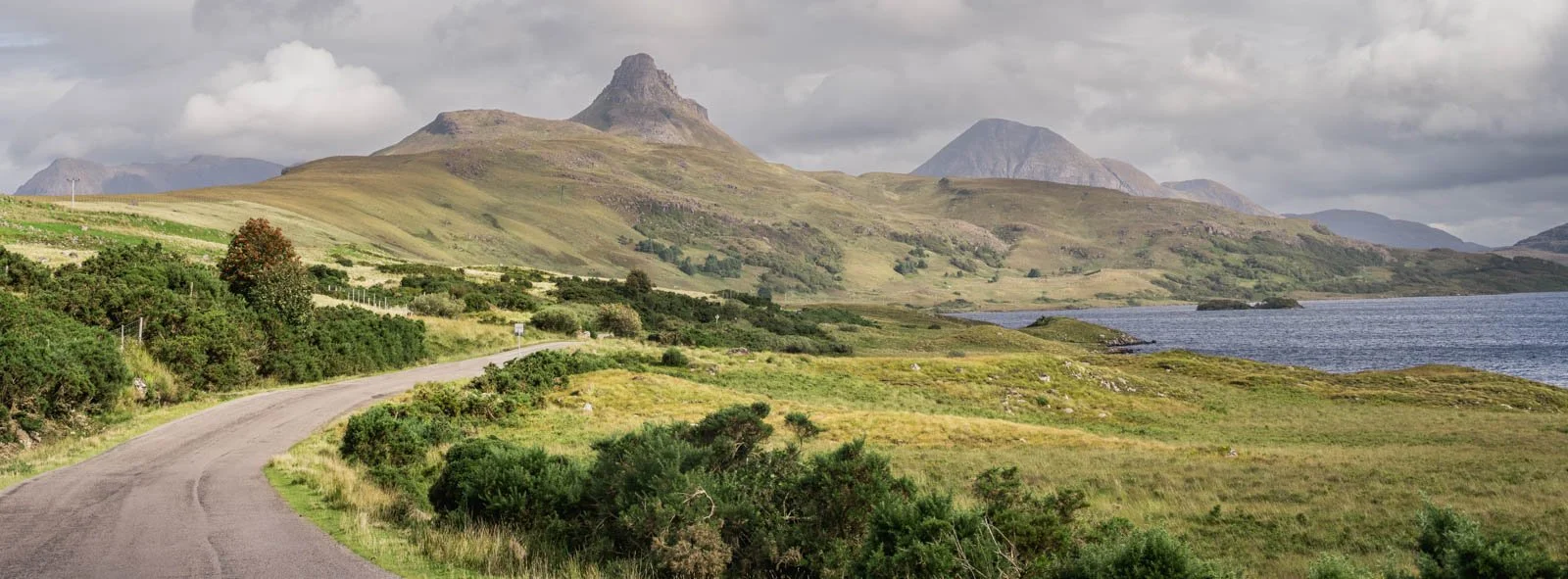 Scenic landscape with a winding road leading towards rolling grassy hills, mountains in the background, and a body of water on the right, under a cloudy sky.