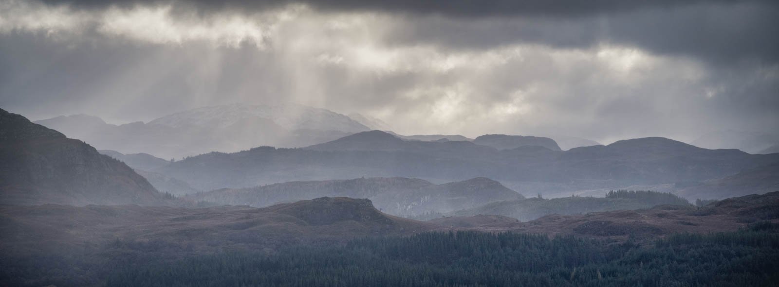 Scenic view of layered mountain ranges under cloudy sky with sunlight breaking through clouds.