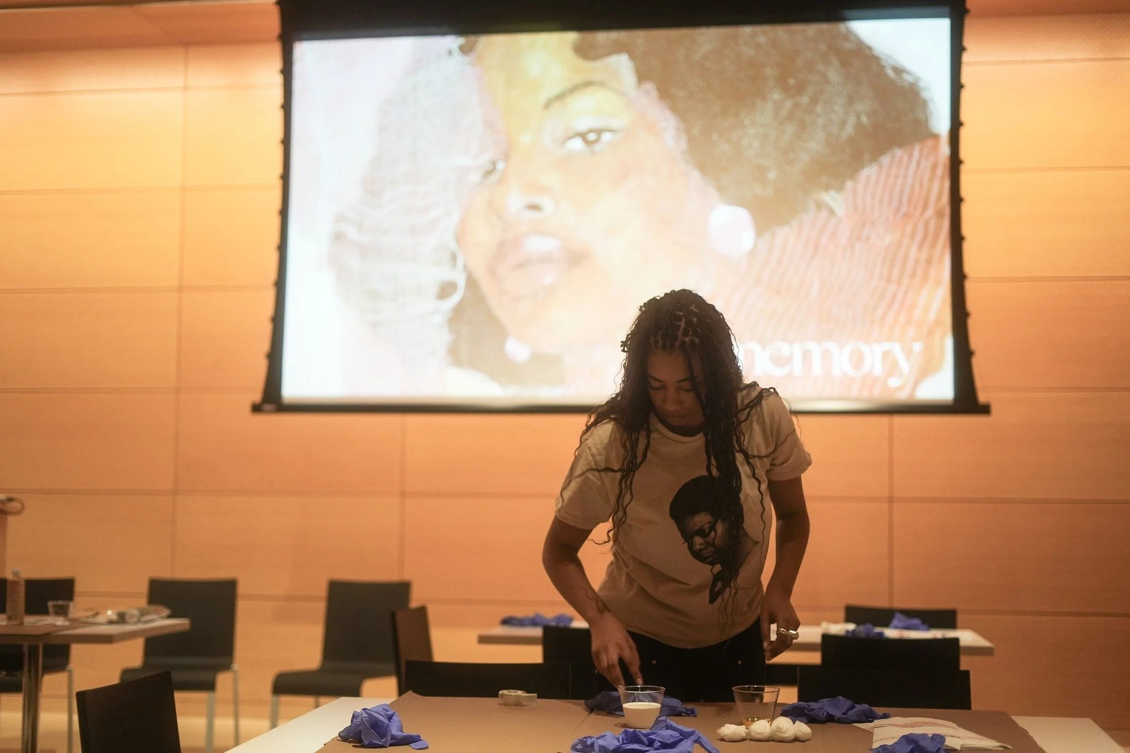 A woman with long braided hair and a graphic t-shirt standing by a table with blue gloves and small objects in a room with a large projected image of a woman's face on the wall behind her.