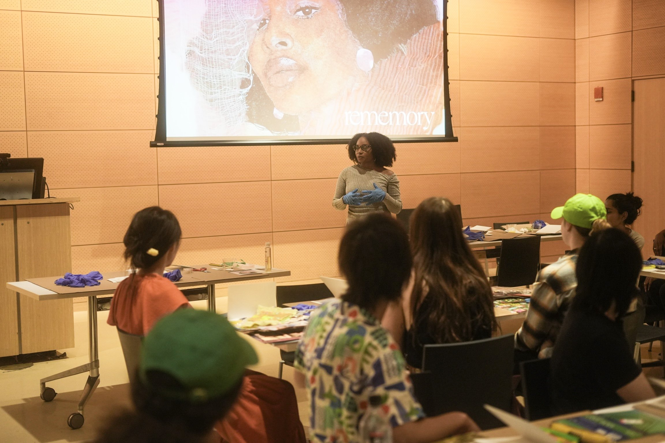 A woman giving a presentation to a group of people in a classroom or conference room with wooden walls, a projector screen, and tables with supplies.