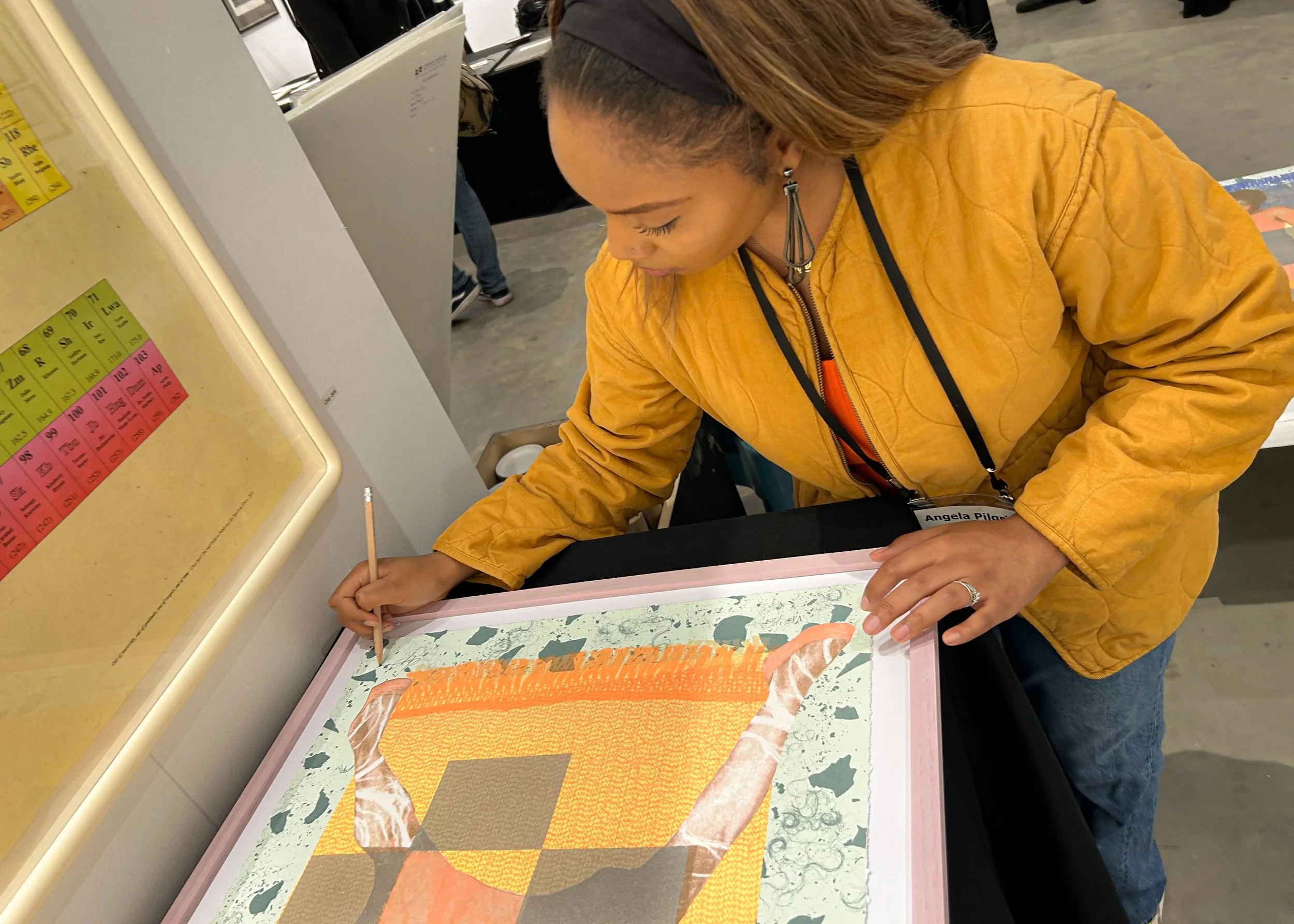 A woman wearing a mustard-colored jacket is signing or drawing on a framed artwork at an exhibition or event.