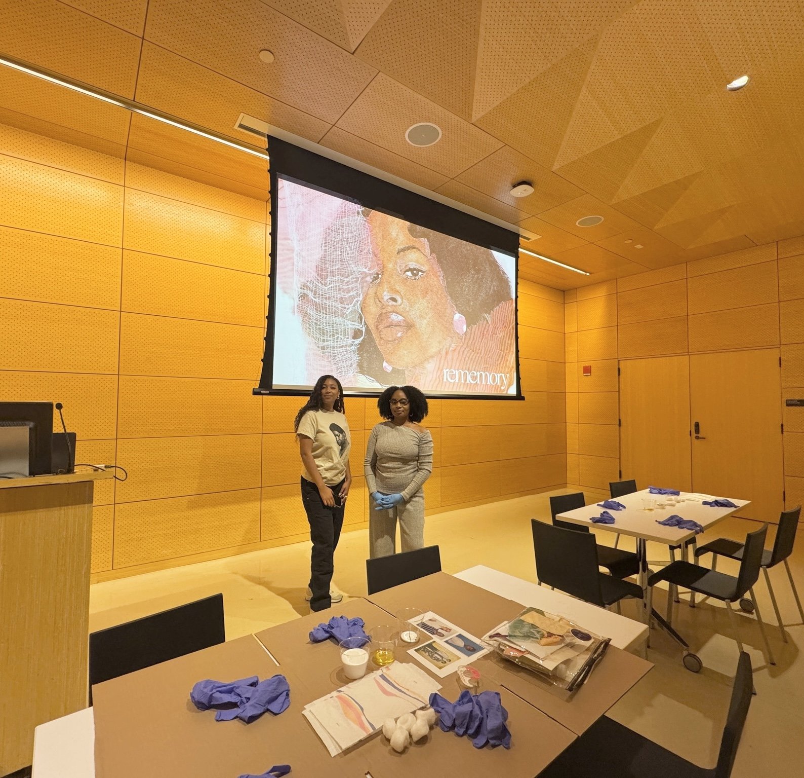 Two women standing in a conference room with a large projected image of a woman's face on the wall behind them. The room has wooden walls and tables with blue gloves and lab materials on them.