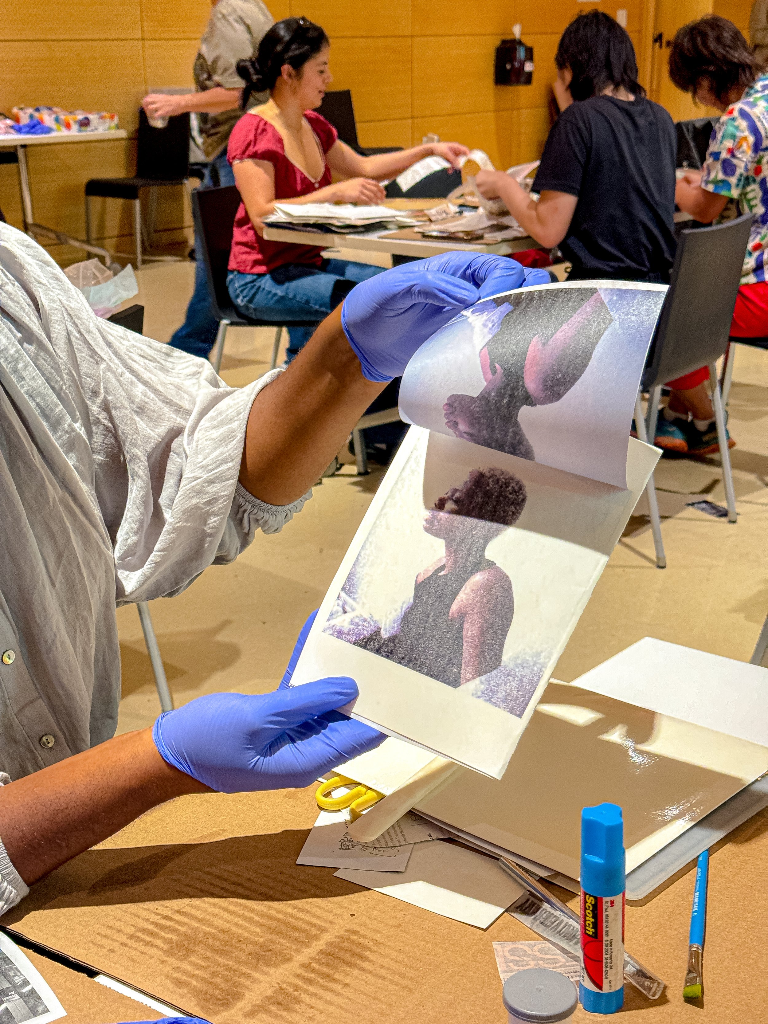 Person wearing blue gloves handling a printed photographic image of a woman with dark hair, wearing a sleeveless top, with other people sitting at tables in the background.