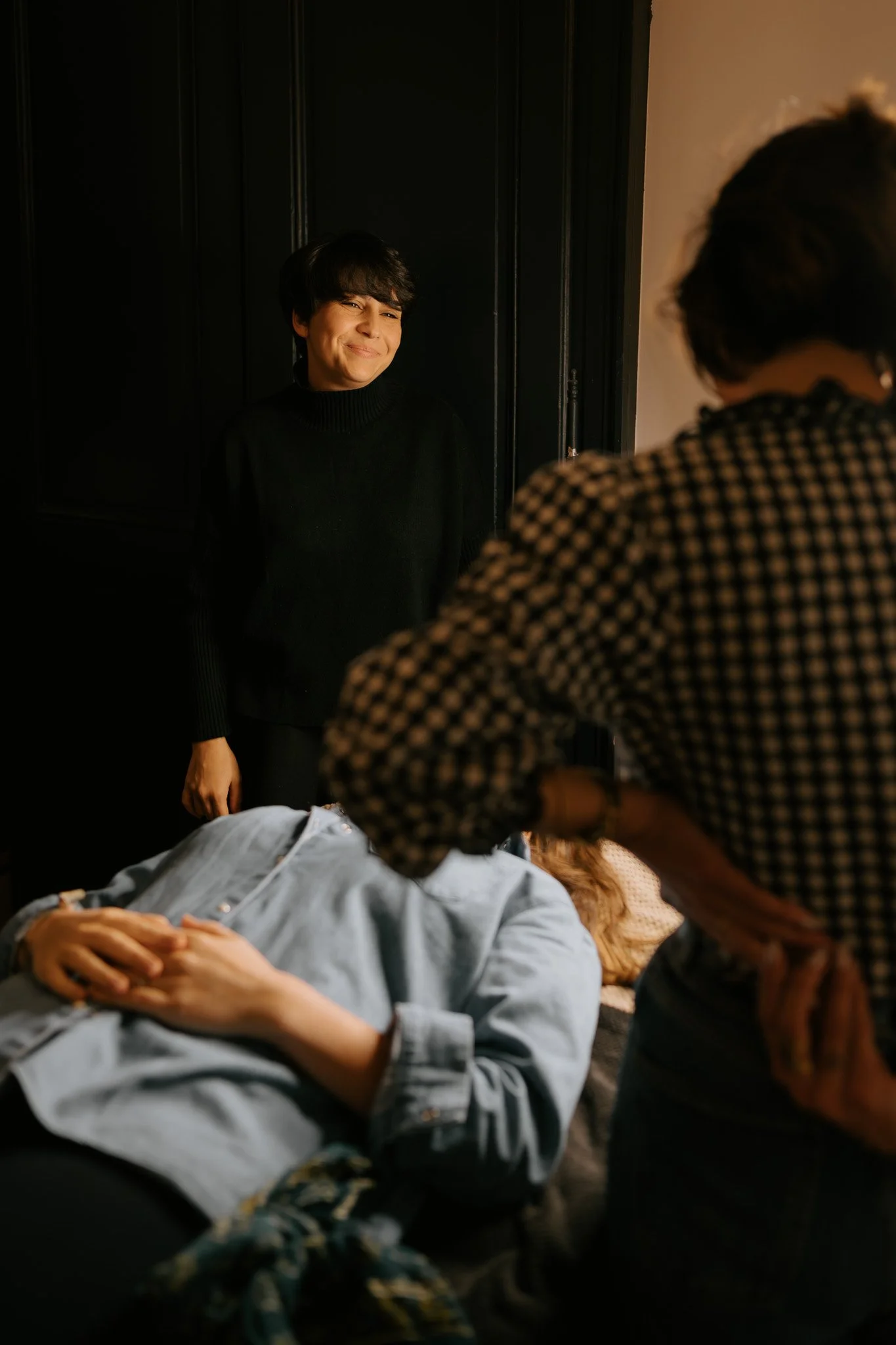 Une jeune femme allongée sur un canapé avec ses mains croisées sur le ventre, assistant à une séance photo avec deux autres femmes, une d'elles face à elle et l'autre de dos, dans un intérieur sombre et chaleureux.