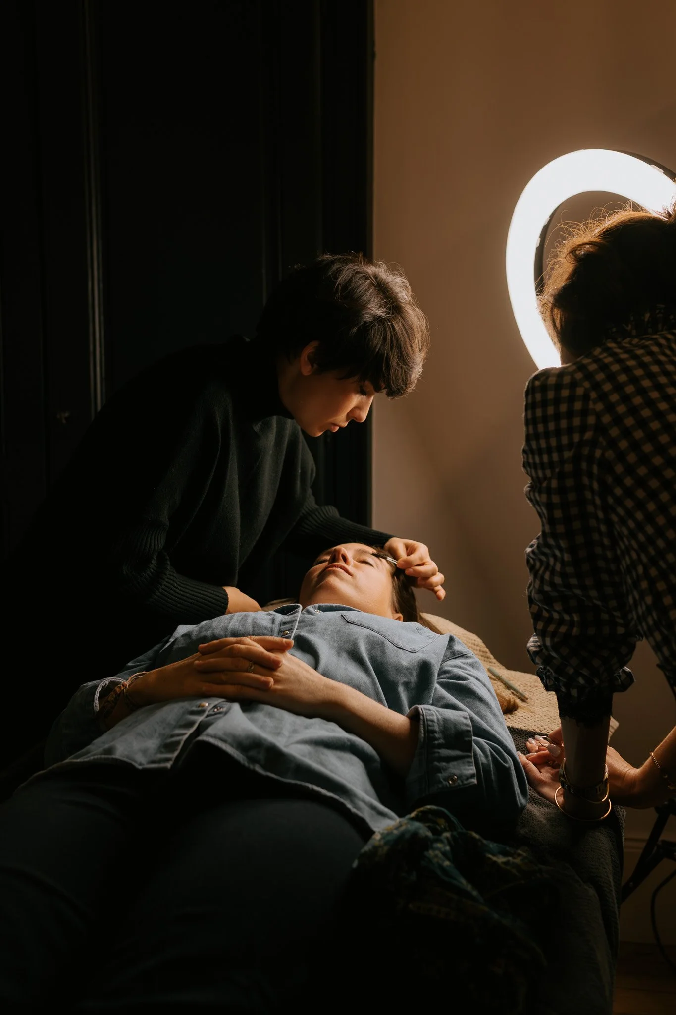 Une personne allongée sur une table, entourée de deux autres personnes, dans un environnement sombre avec un éclairage lumineux. Il semble que cette personne reçoit des soins ou un traitement facial.