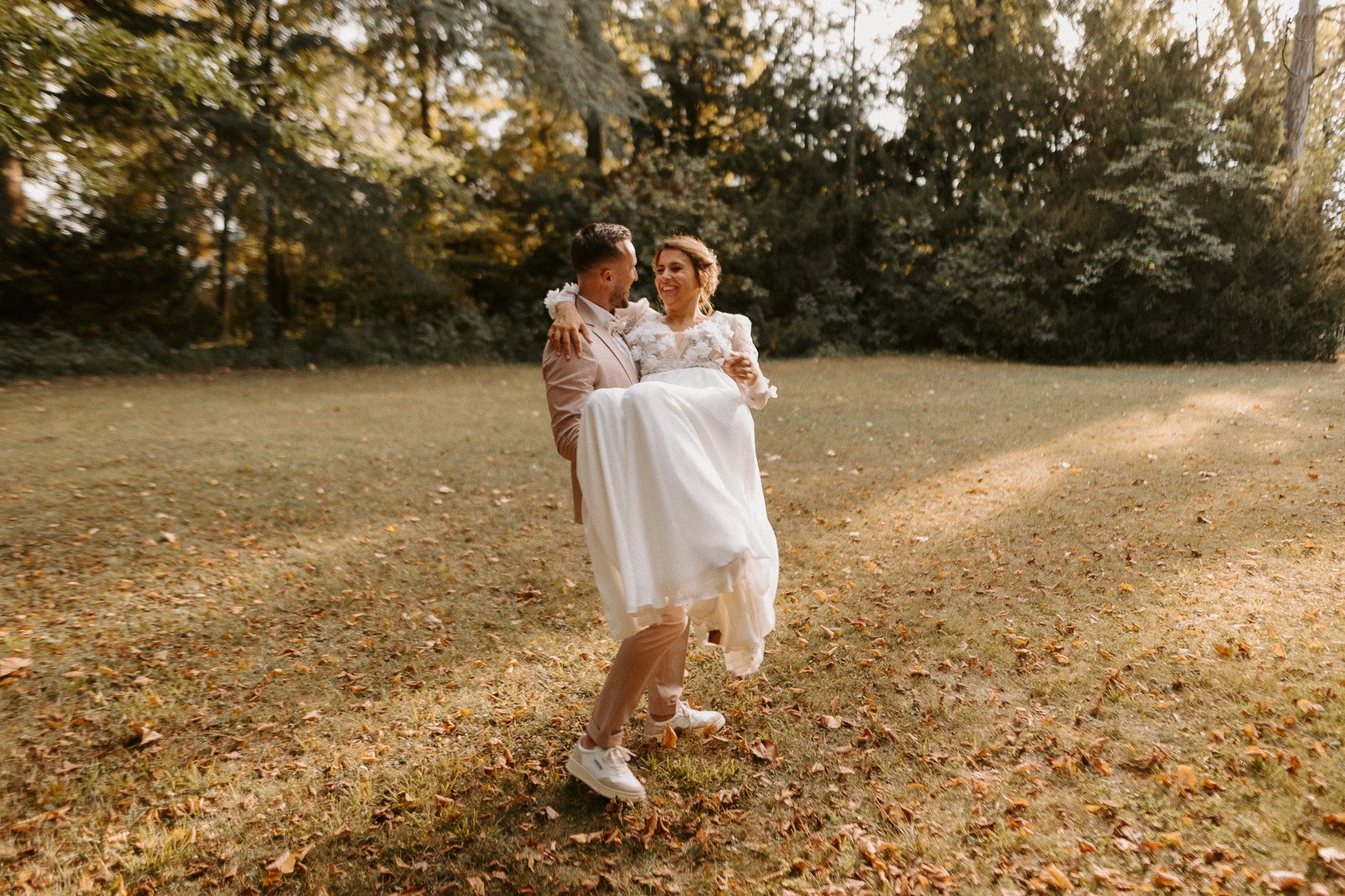 Un couple de mariés dans un parc, le marié porte la mariée dans ses bras, tous deux souriants, entourés d'arbres aux feuilles d'automne.