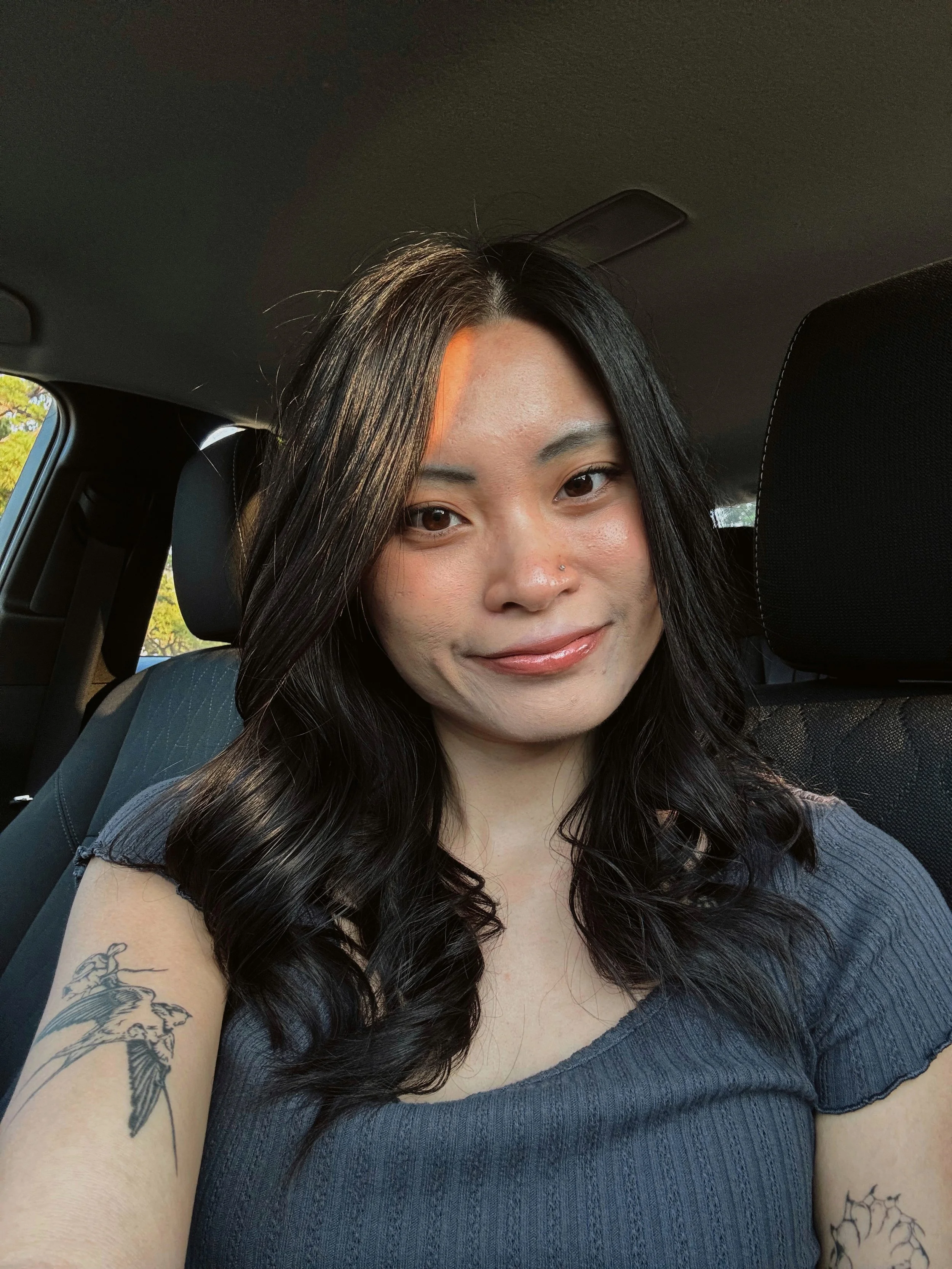 A woman with long black curly hair, wearing a gray sleeveless top, looking directly at the camera with a neutral expression, in a dimly lit room.