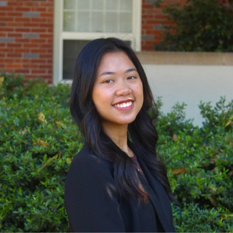 Close-up of a young man smiling, wearing glasses and a gray jacket against a dark background.