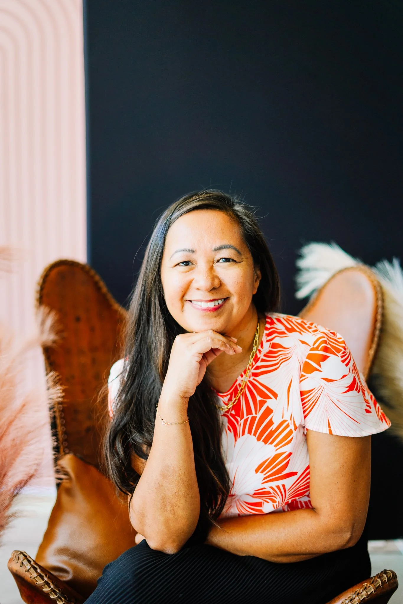 A woman with long dark hair and a bright smile, wearing a white and orange patterned blouse, sitting in a stylish chair with a dark background.