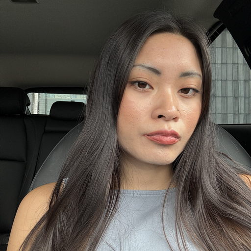 A woman with long black curly hair, wearing a gray sleeveless top, looking directly at the camera with a neutral expression, in a dimly lit room.