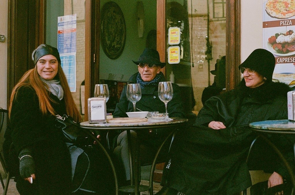 Three people sitting at an outdoor cafe table, dressed in winter clothing, with empty wine glasses and menu visible.