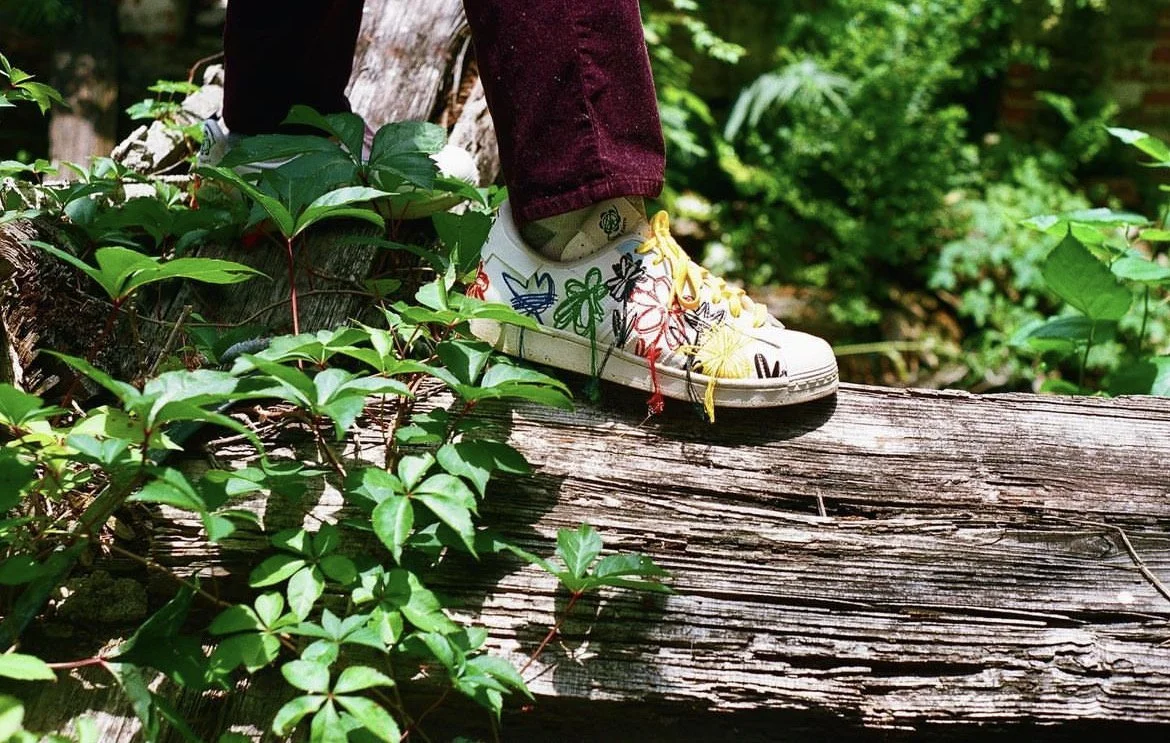 Person wearing floral embroidered sneakers standing on a fallen tree surrounded by greenery.