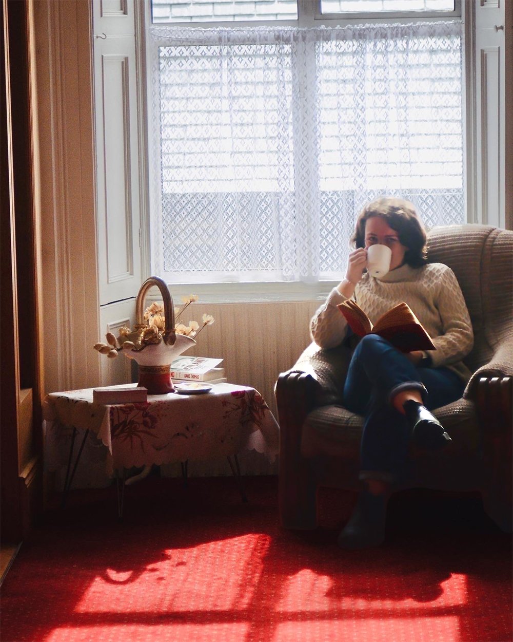 Person sitting in a cozy chair by a window, drinking from a mug and reading a book, with a small table and decorative items nearby, sunlight streaming in.