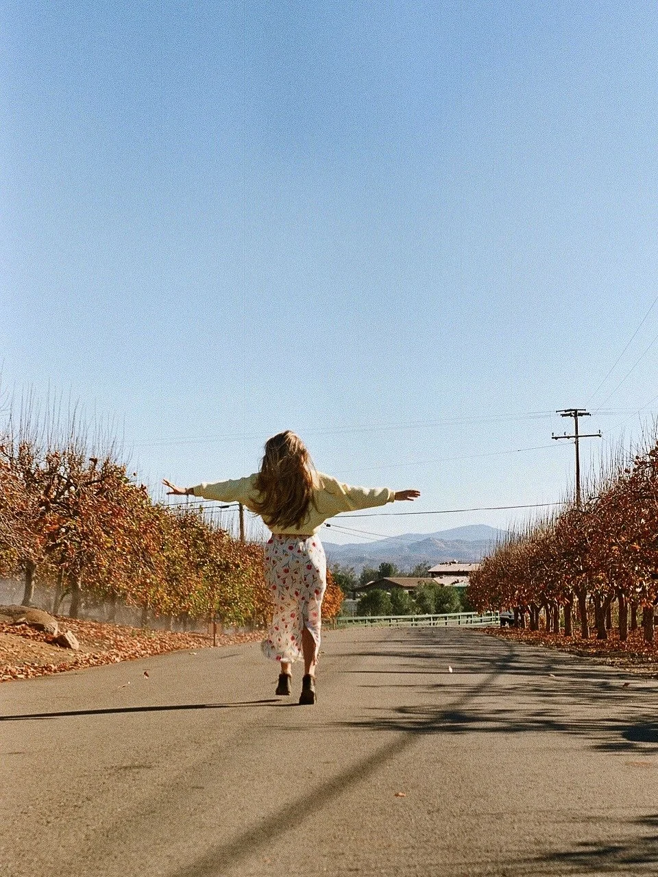 A woman with long hair walks joyfully down a road lined with autumn trees, arms spread wide. She is wearing a light-colored sweater and a floral skirt. The scene is set in a rural area with clear blue skies and mountains in the background.
