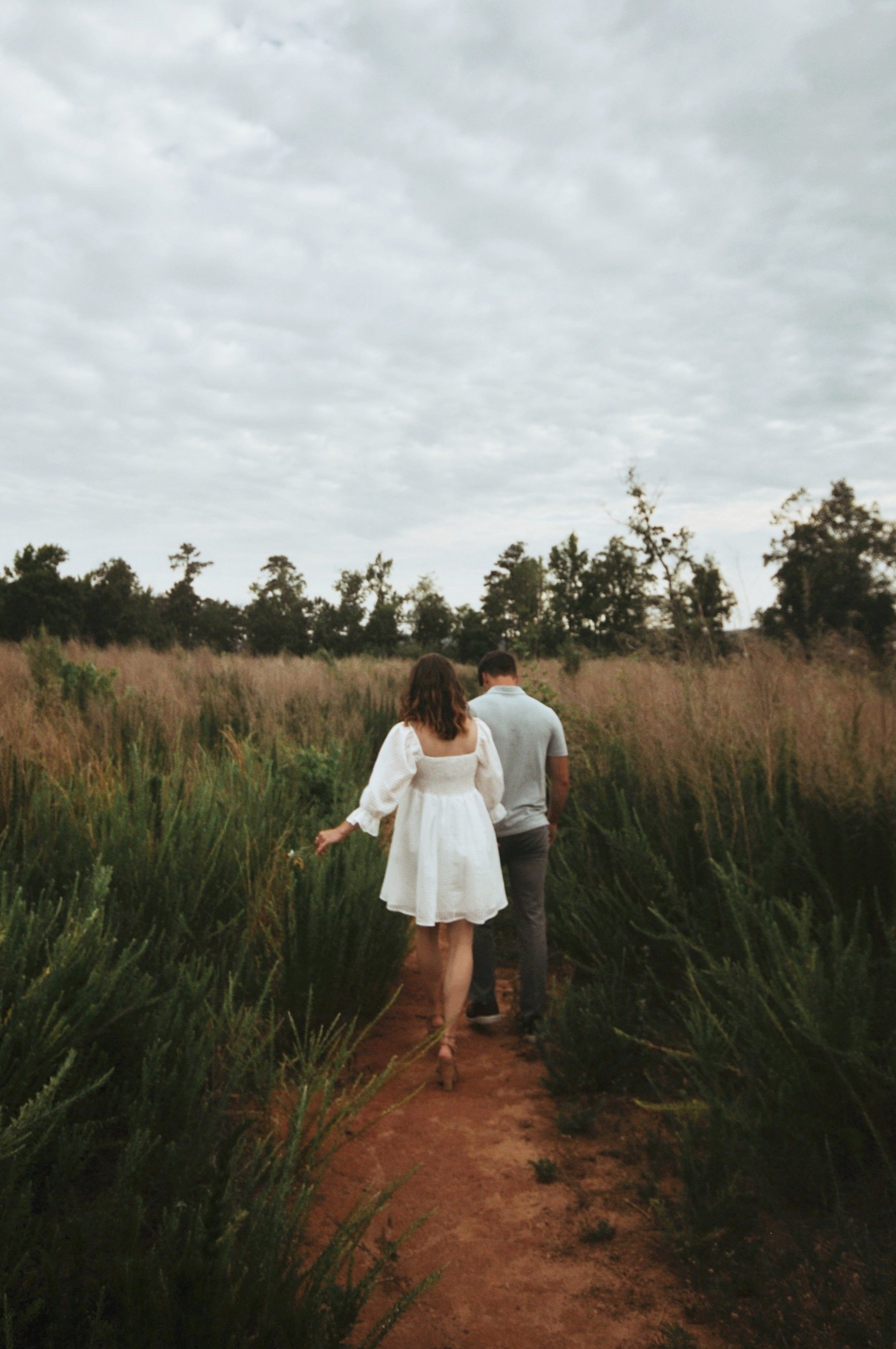 Couple walking on a dirt path through a meadow with tall grass and trees in the background under a cloudy sky.