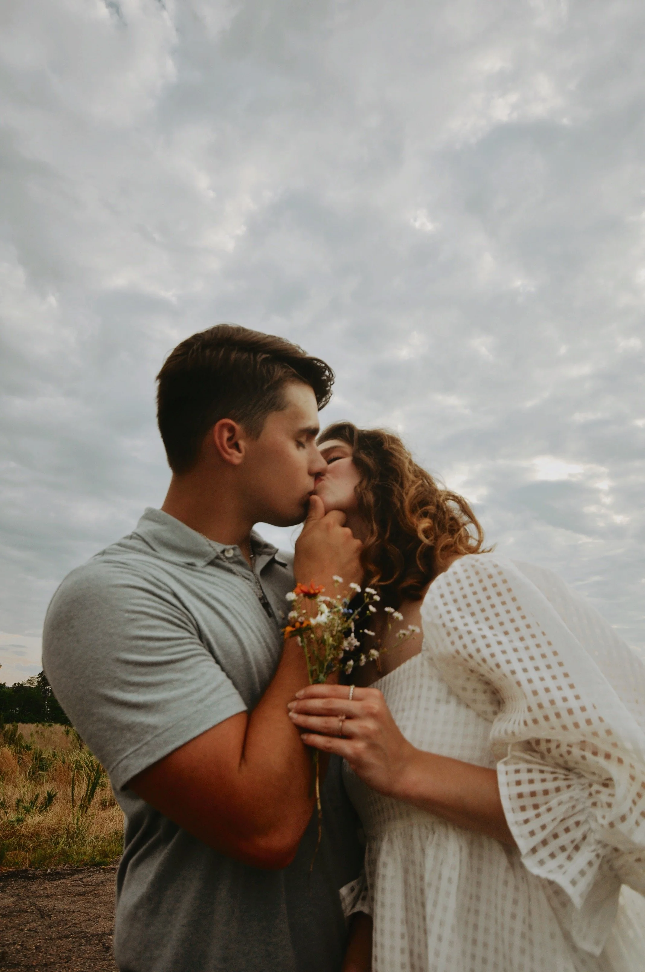 Couple kissing outdoors with a cloudy sky, holding a small bouquet of wildflowers.
