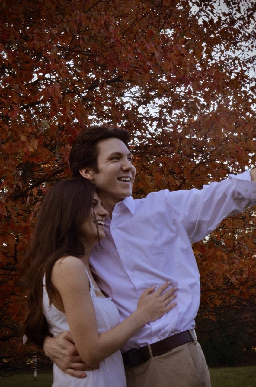 A couple embracing and smiling in front of autumn foliage.