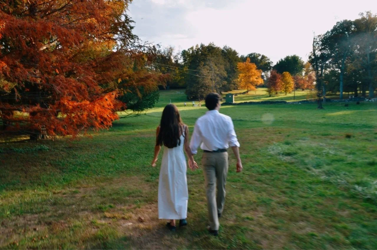 A couple walking hand in hand through a grassy field with vibrant autumn trees in the background.