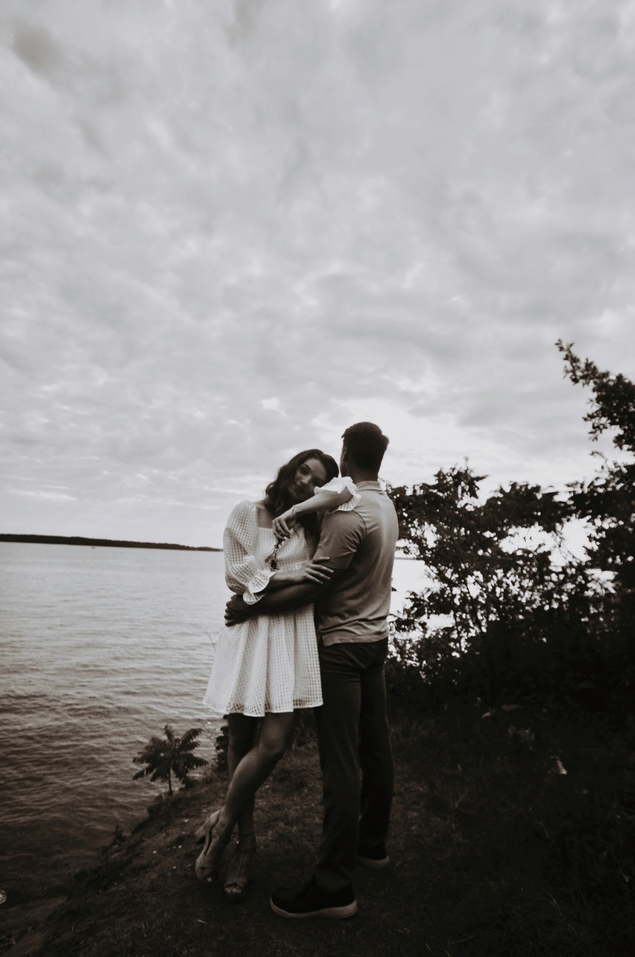 A couple embracing by a lake with cloudy skies. The woman is wearing a white dress and leaning against the man, who has his back to the camera. Trees and water are in the background.