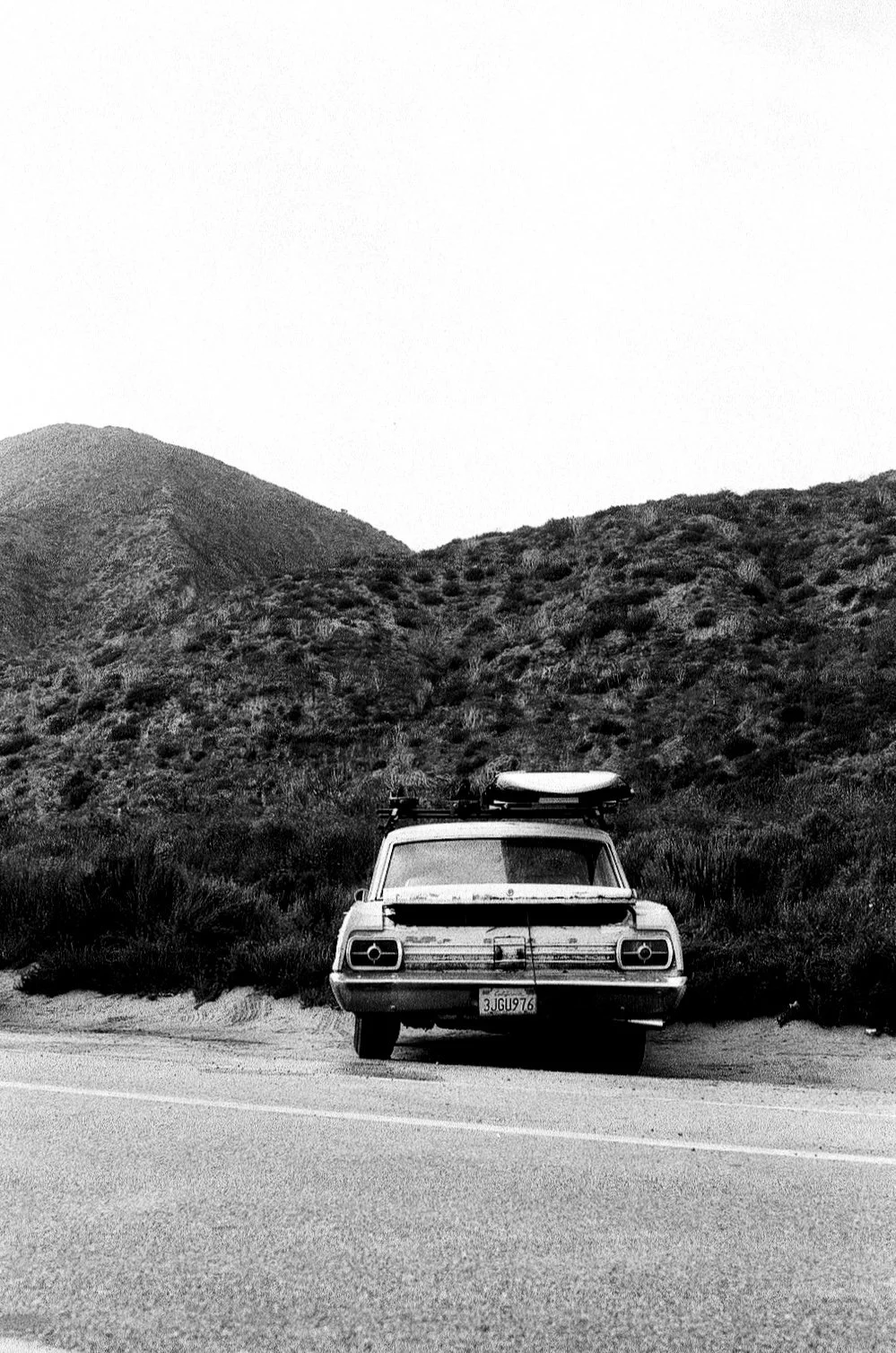 Black and white photo of a vintage car with a surfboard on top parked by the roadside, with hills in the background.