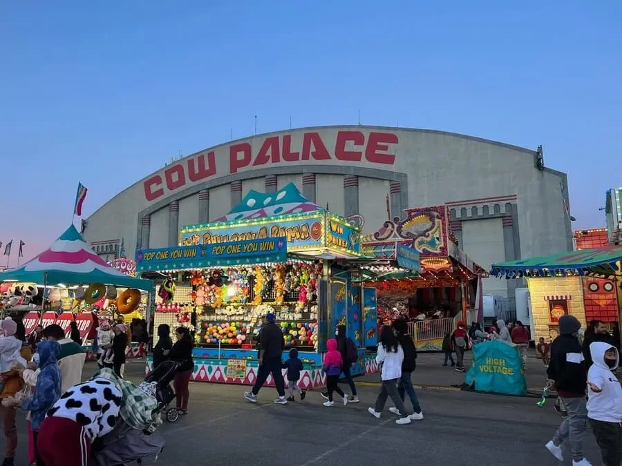 View of a fairground with colorful rides and game booths in front of a large building labeled 'COW PALACE'. People are walking around, some pushing strollers and engaging with the attractions.