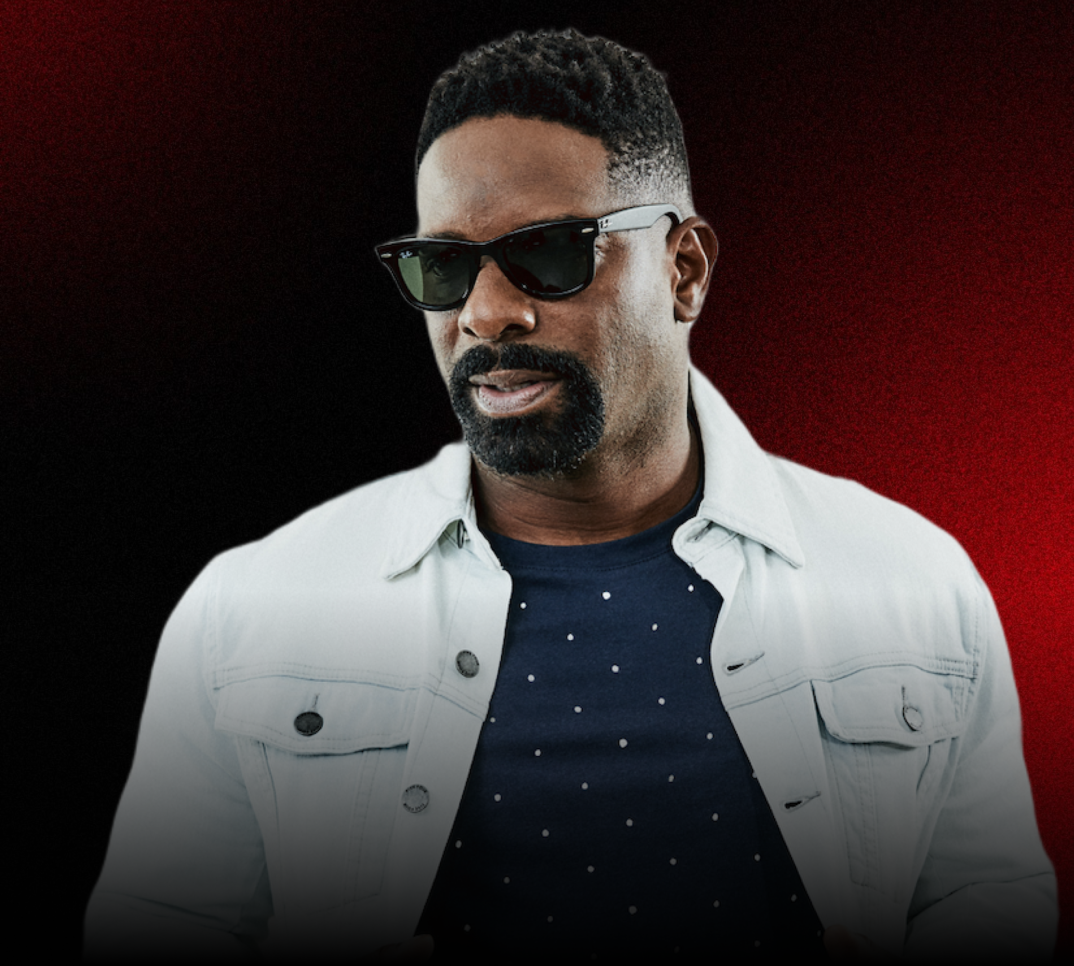 Man wearing black sunglasses, a white denim jacket, and a navy shirt with white polka dots, standing against a black and red background.