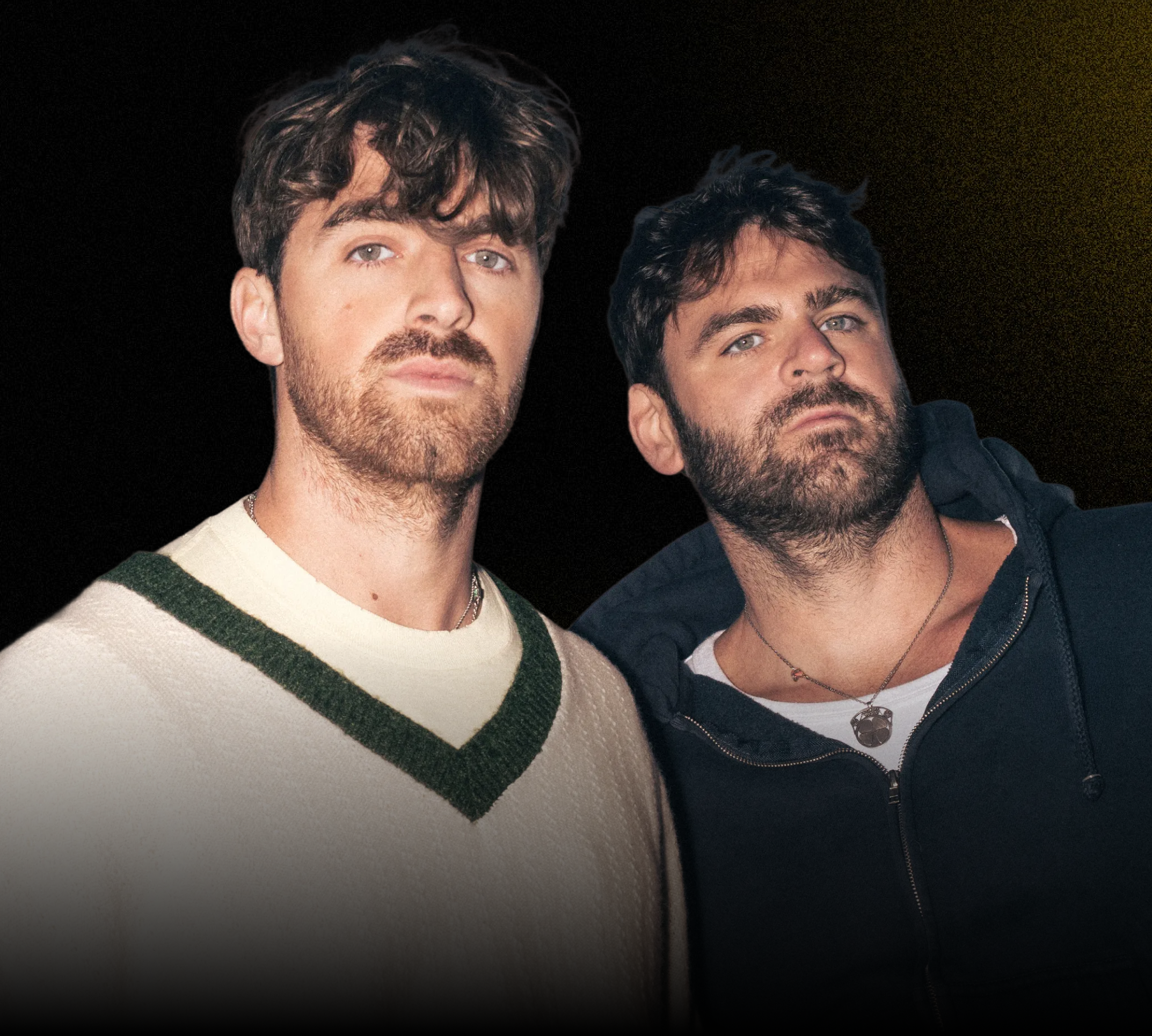 Two men with dark hair and beards standing close together against a dark background.