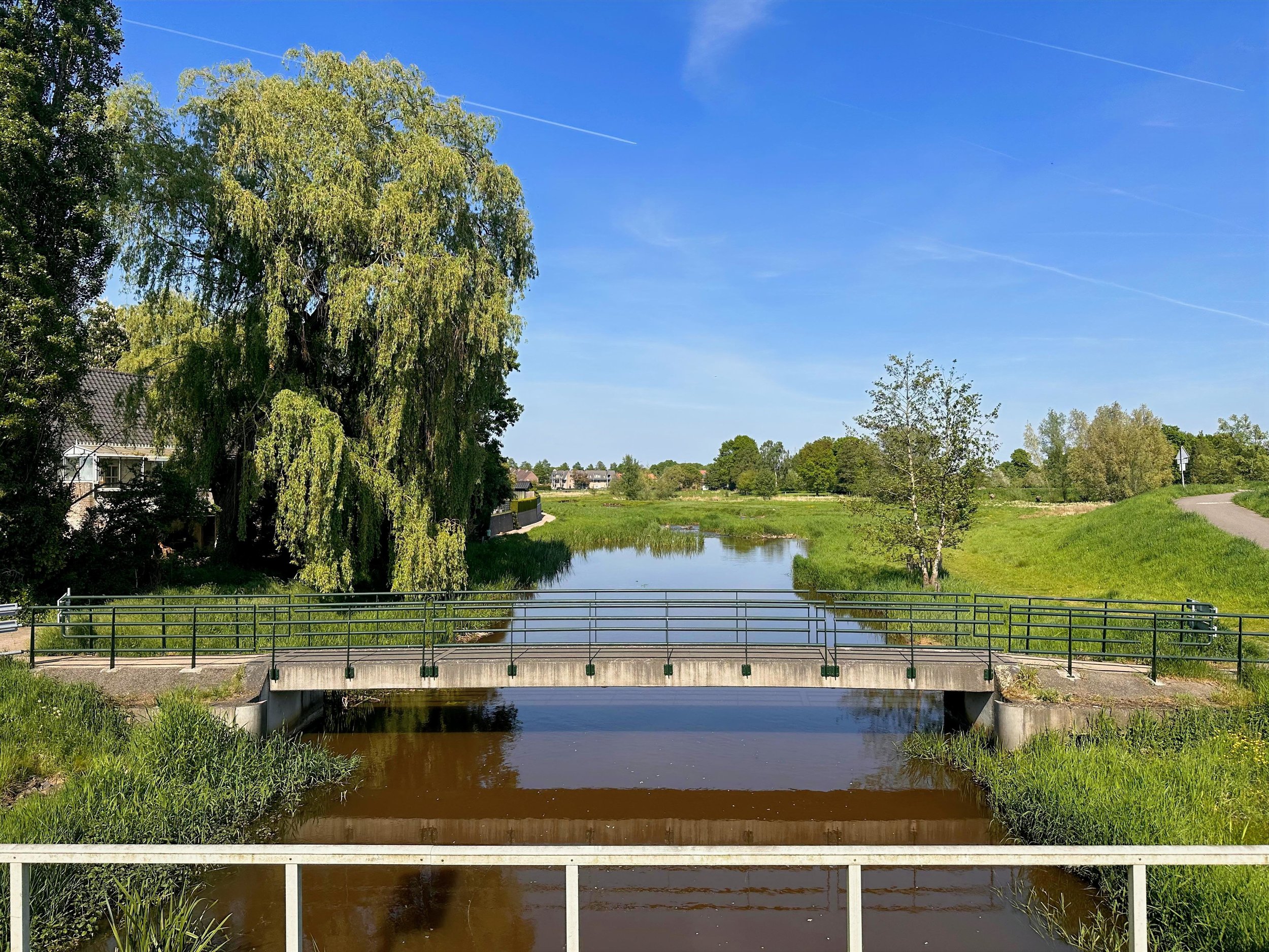 Een kleine rivier met een brug erover, omringd door groene bomen en een open veld, onder een blauwe lucht met enkele contrails.