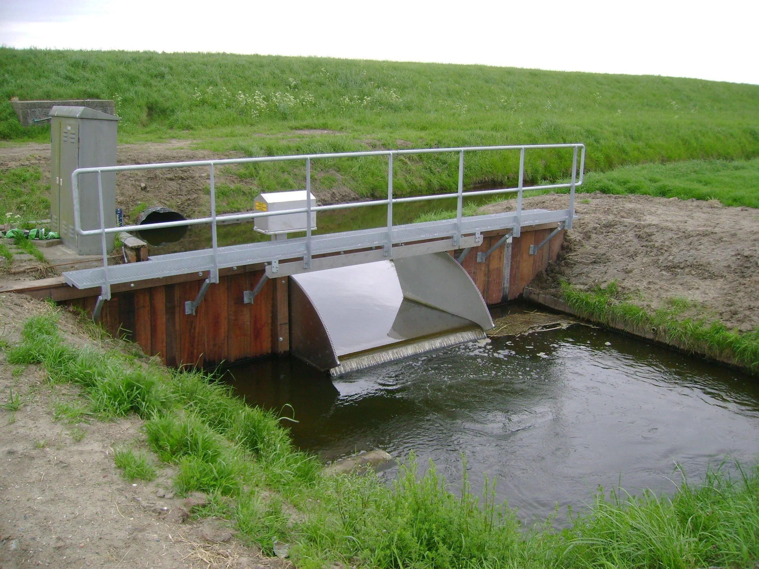 Een klein watergemaal met een metalen brug over een waterloop, omgeven door groene graslanden en een heuvel in de achtergrond.