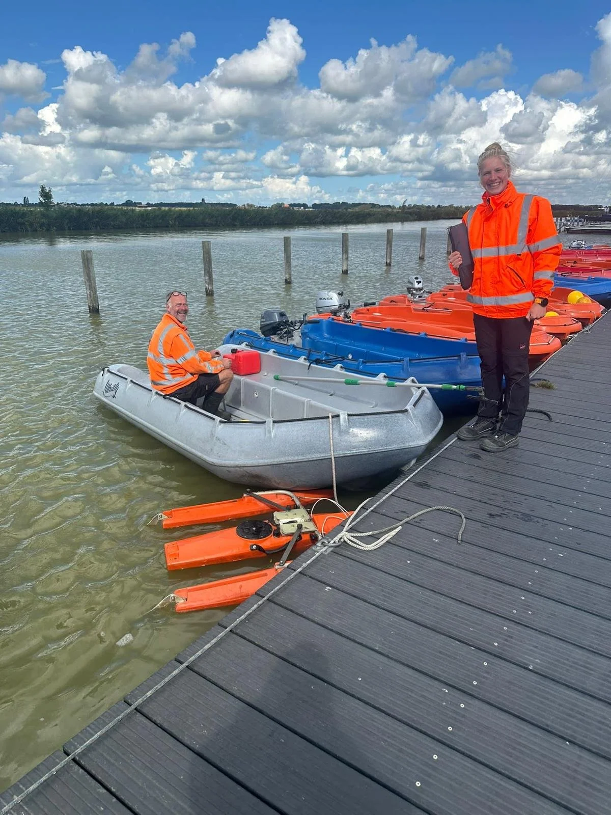 Twee mensen in oranje veiligheidsjassen bij een boot aan de kade, klaar voor vaartocht, onder een bewolkte hemel.