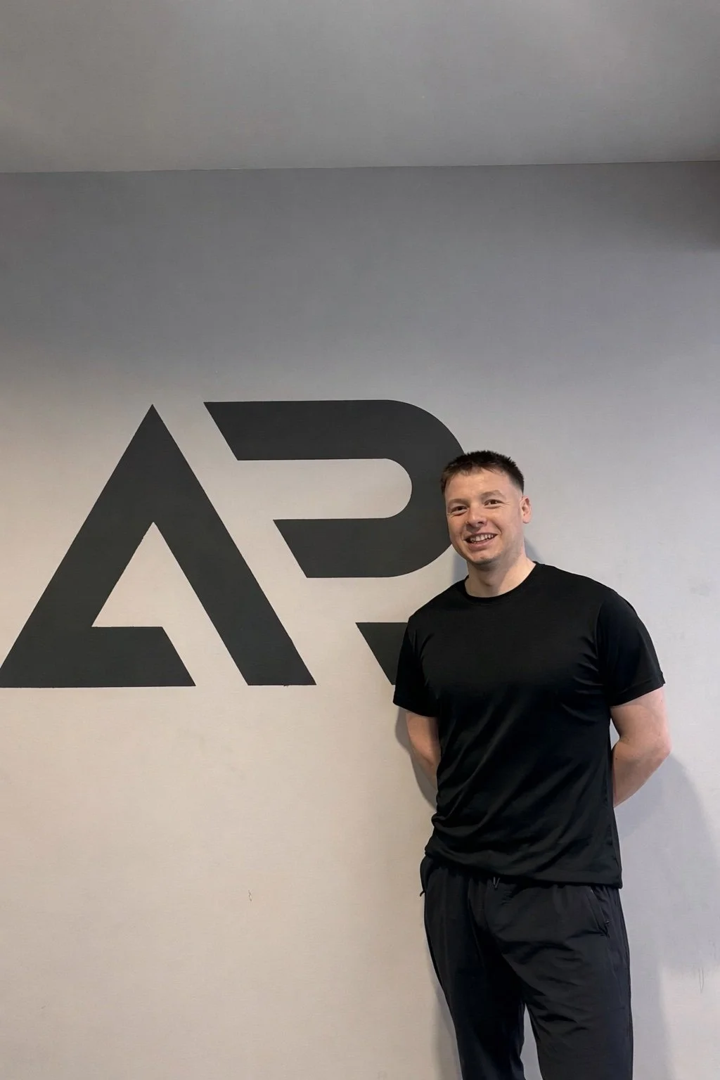 A man in a black t-shirt and black pants standing in front of a wall with a large black logo.