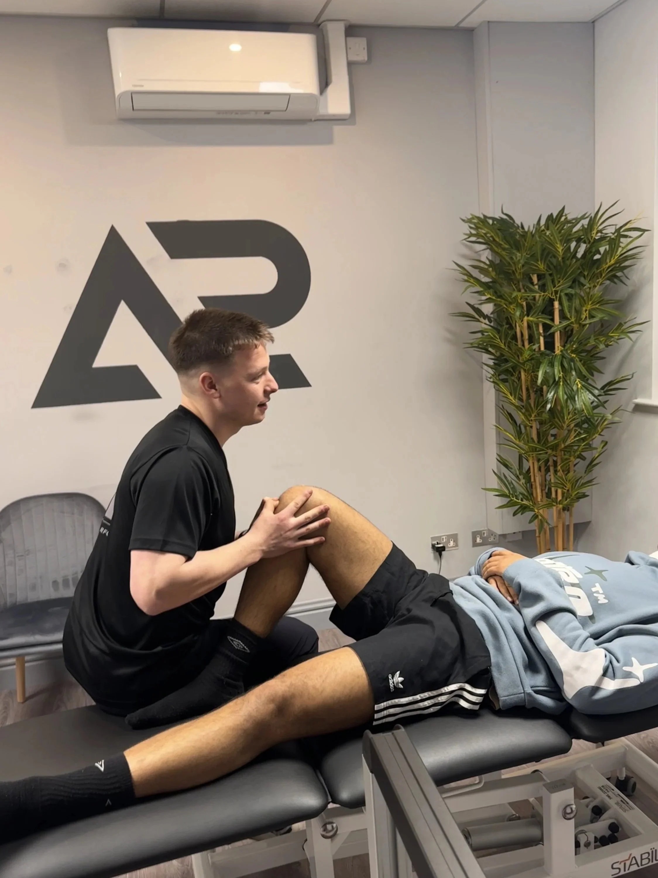 A physical therapist or trainer is performing a treatment or exercise on a male patient who is lying on a therapy table, holding his knee while the therapist supports his leg. They are in a clinical setting with a large logo on the wall, an air conditioning unit, a tall plant, and various electrical outlets.