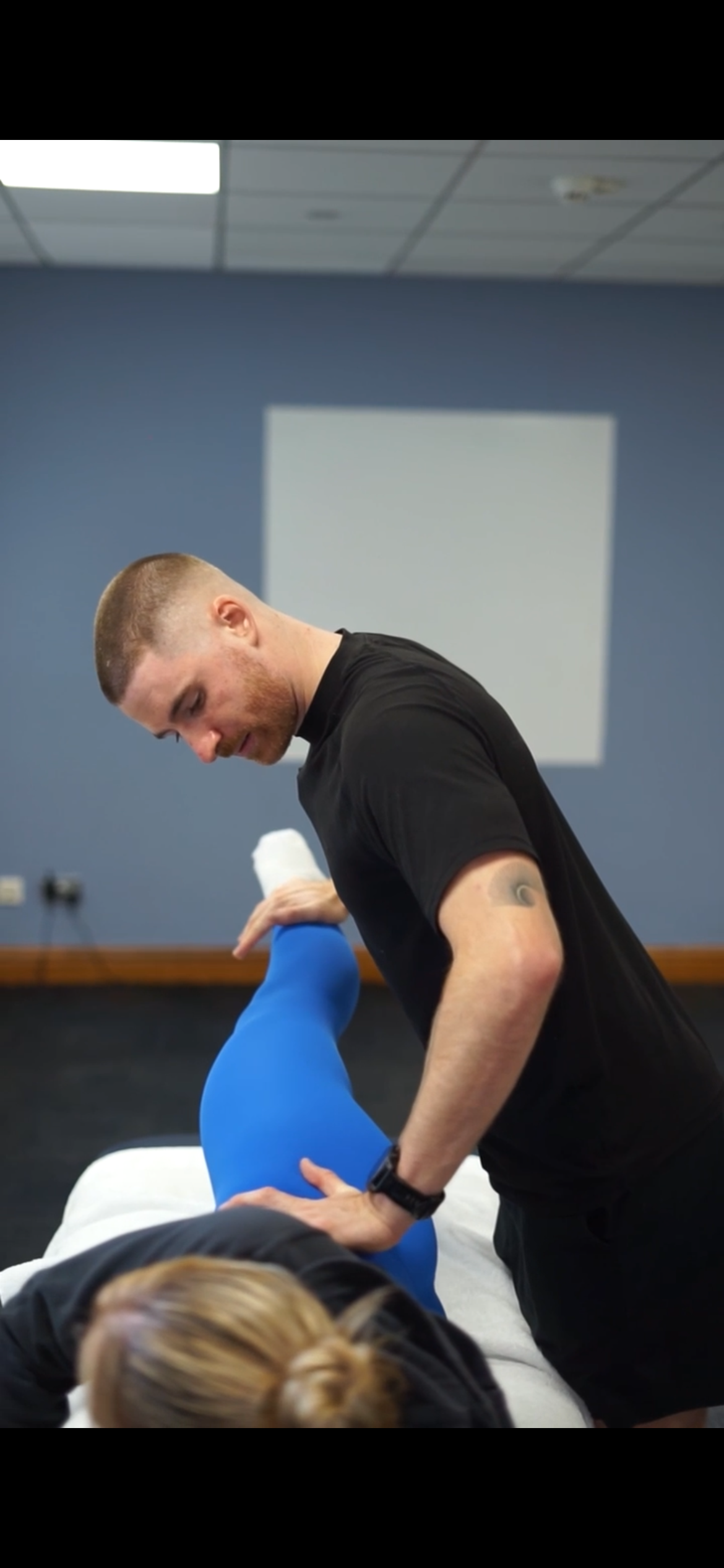 A man appears to be a physical therapist or trainer, assisting a woman with her leg in a gym or therapy room. The room has blue walls and a whiteboard or picture on the wall.